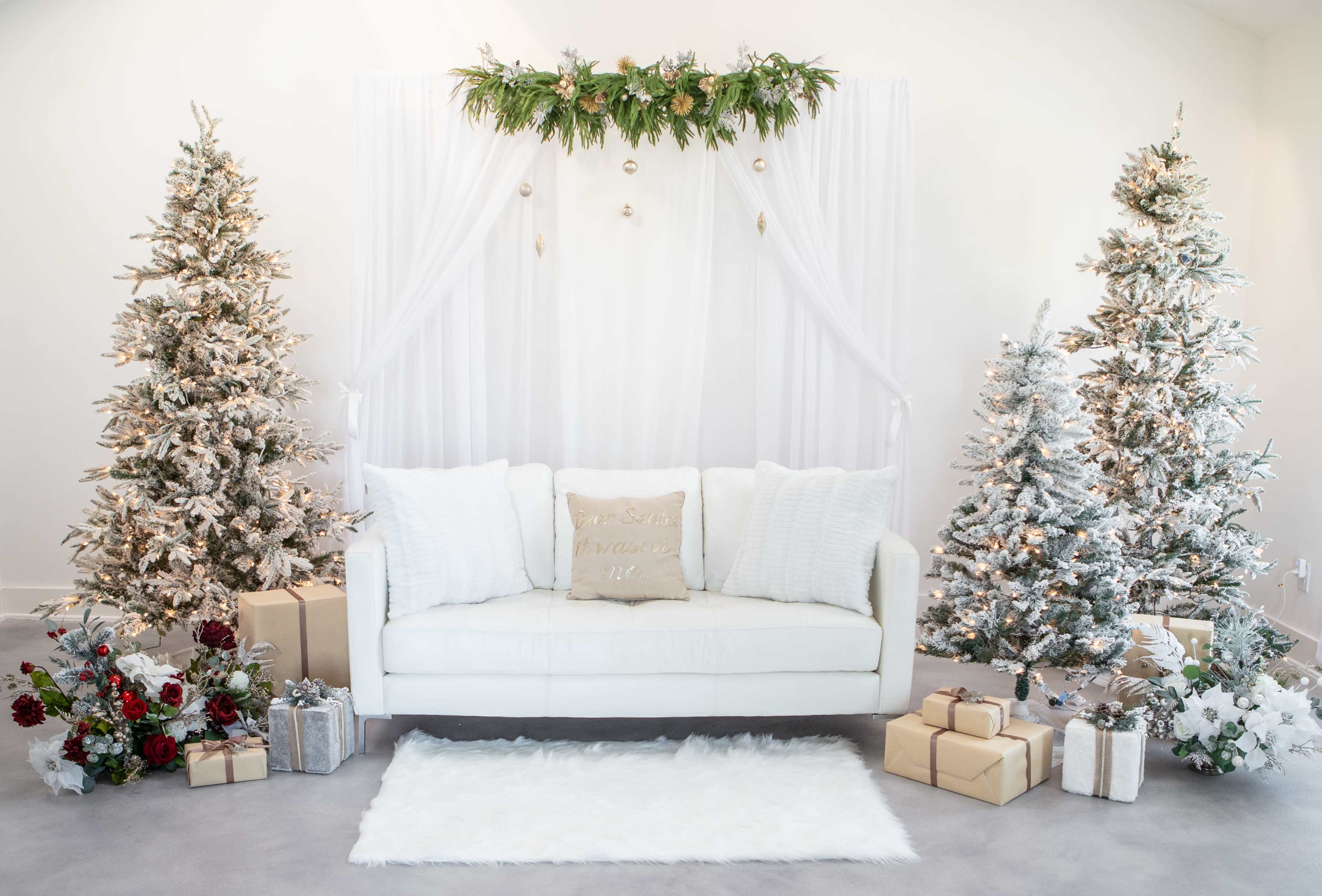 The scene features a white sofa adorned with decorative pillows, flanked by two frosted Christmas trees and surrounded by wrapped presents, all set against a backdrop of draped white fabric and greenery.