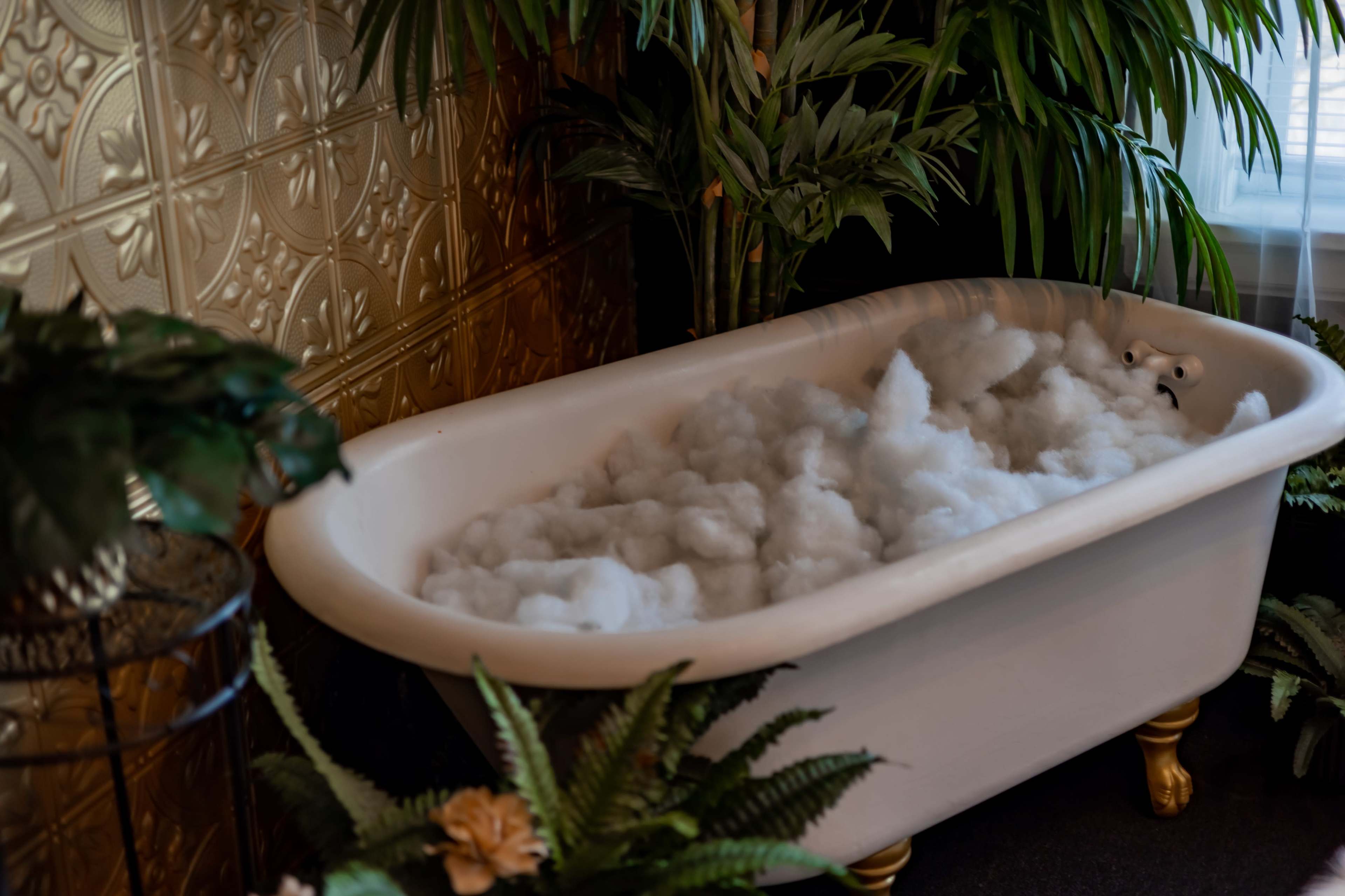 A vintage clawfoot bathtub filled with bubbles sits among lush green plants against a patterned gold wall.