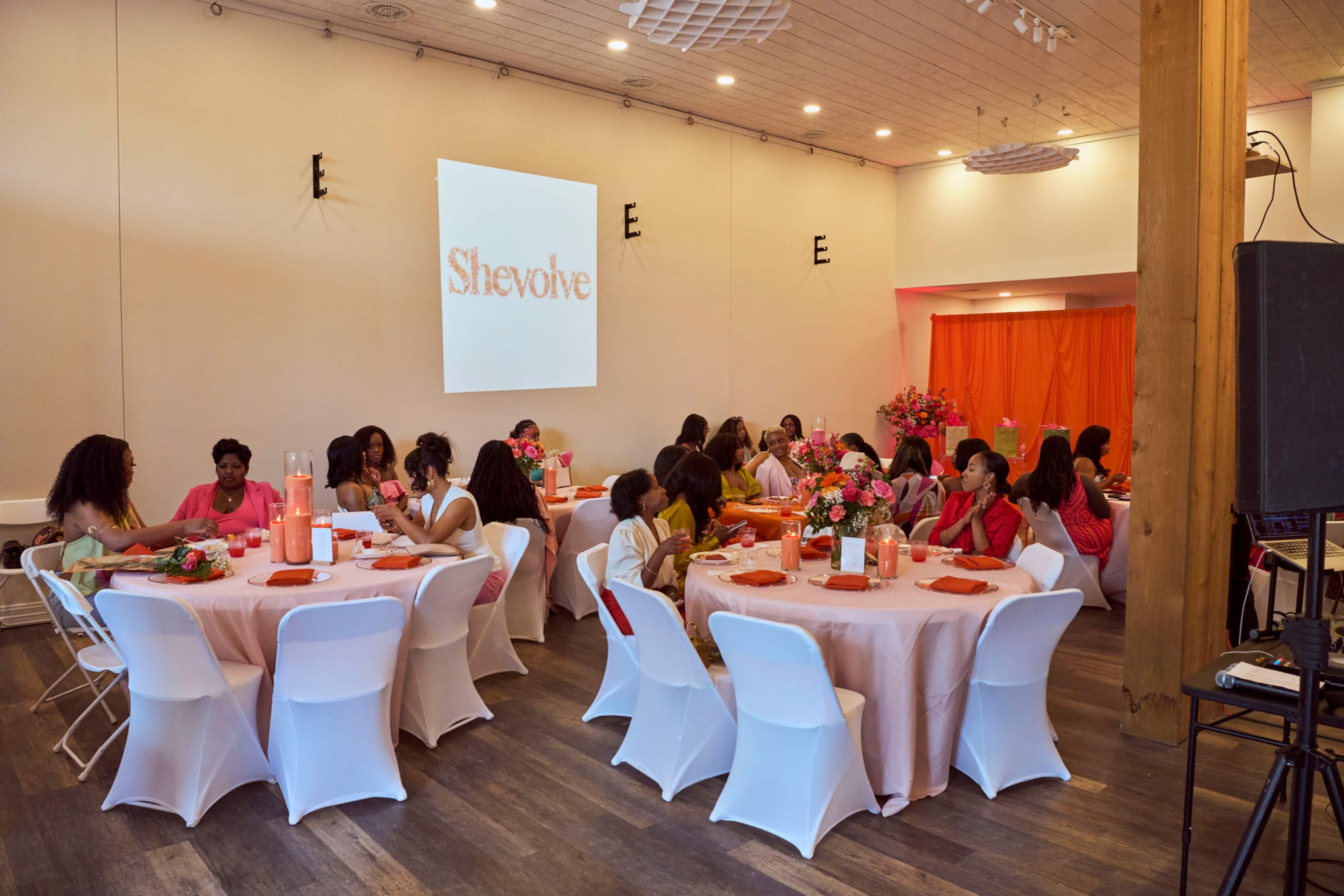 A group of women gather around tables at an event featuring orange decorations and a presentation screen displaying the word "Shevolve."