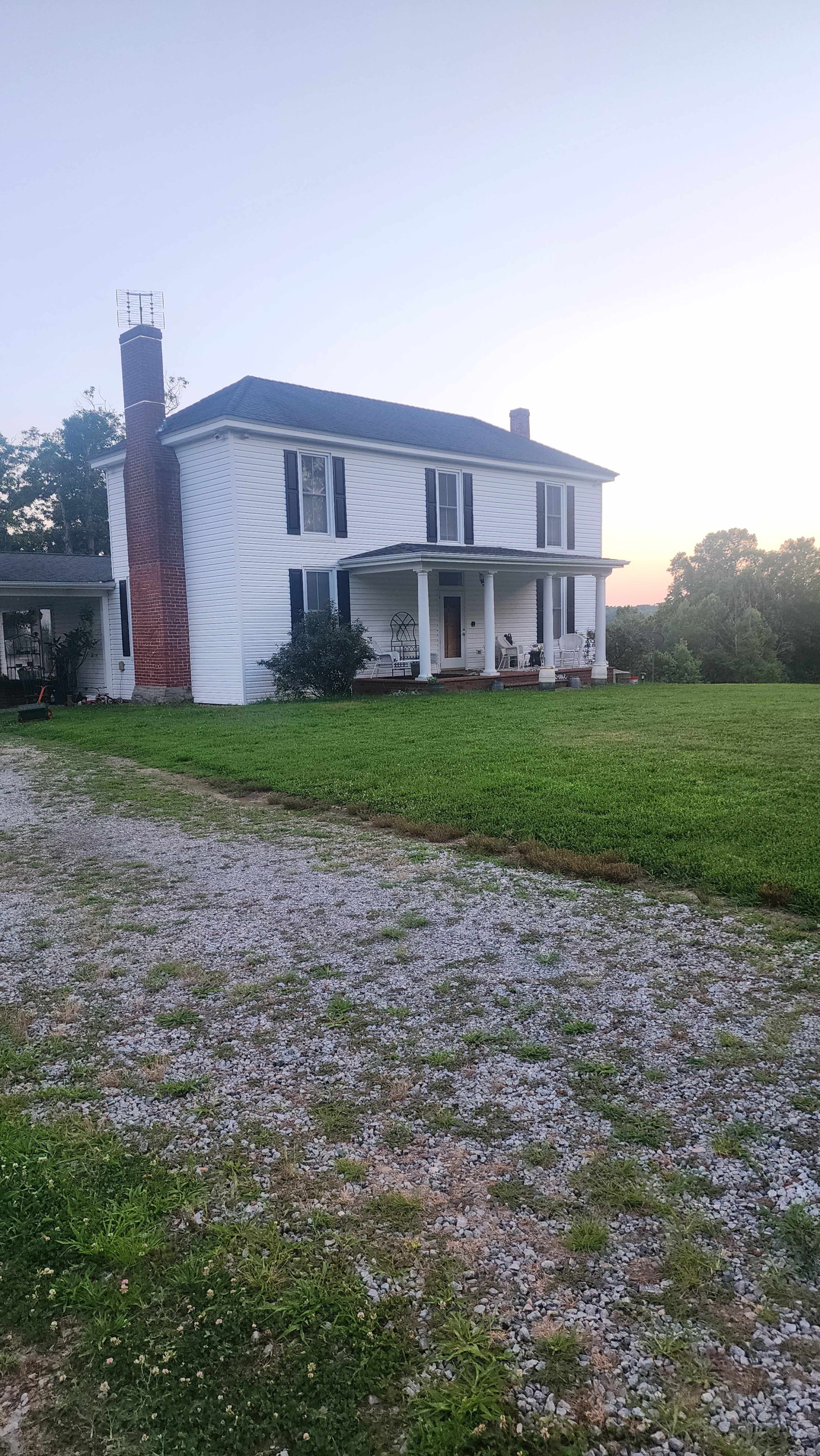 A two-story white house with a gray roof sits at the end of a gravel driveway, surrounded by a grassy lawn.