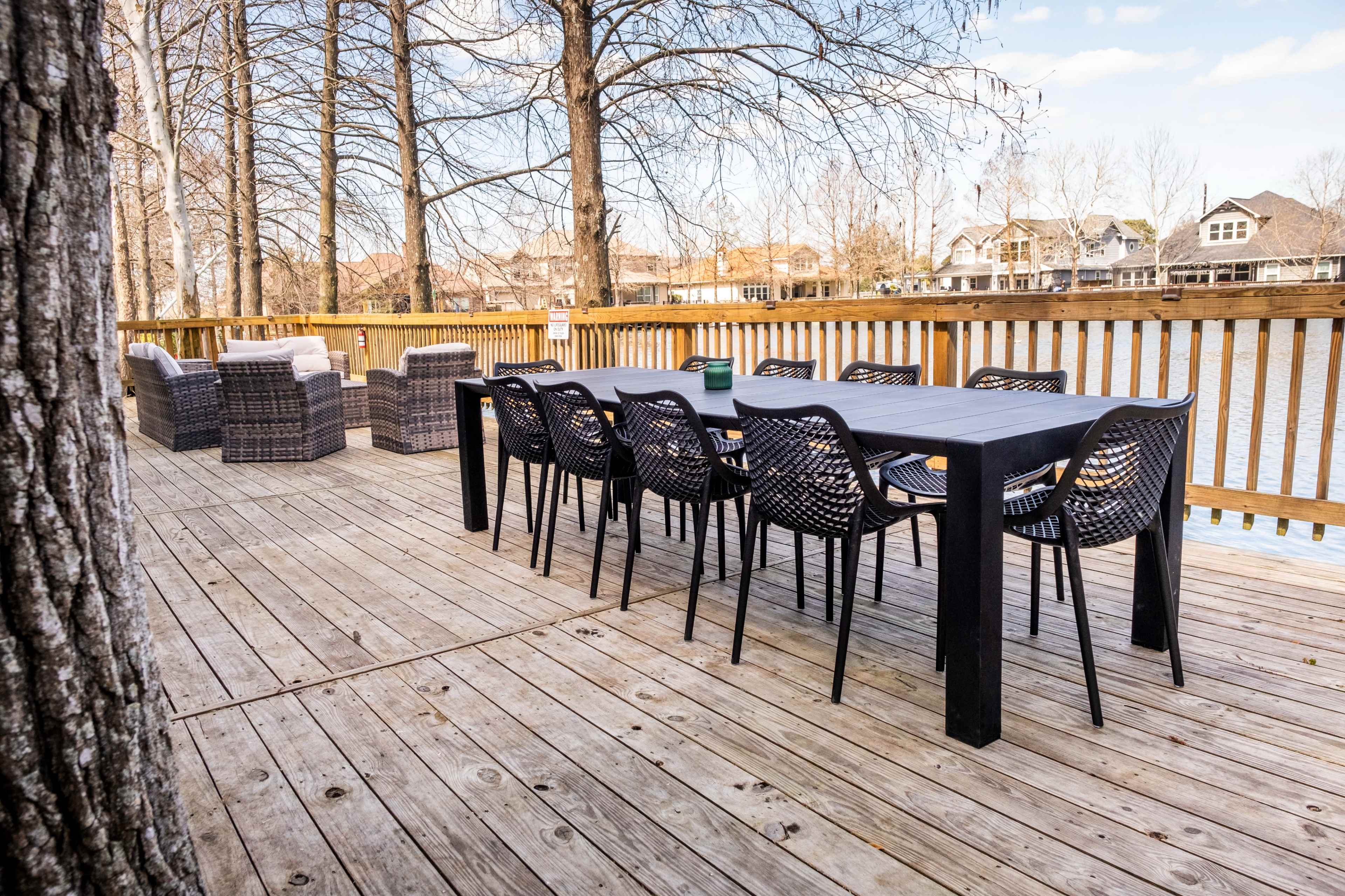 A long black table surrounded by matching chairs is set on a wooden deck near a lake, with wicker seating in the background.