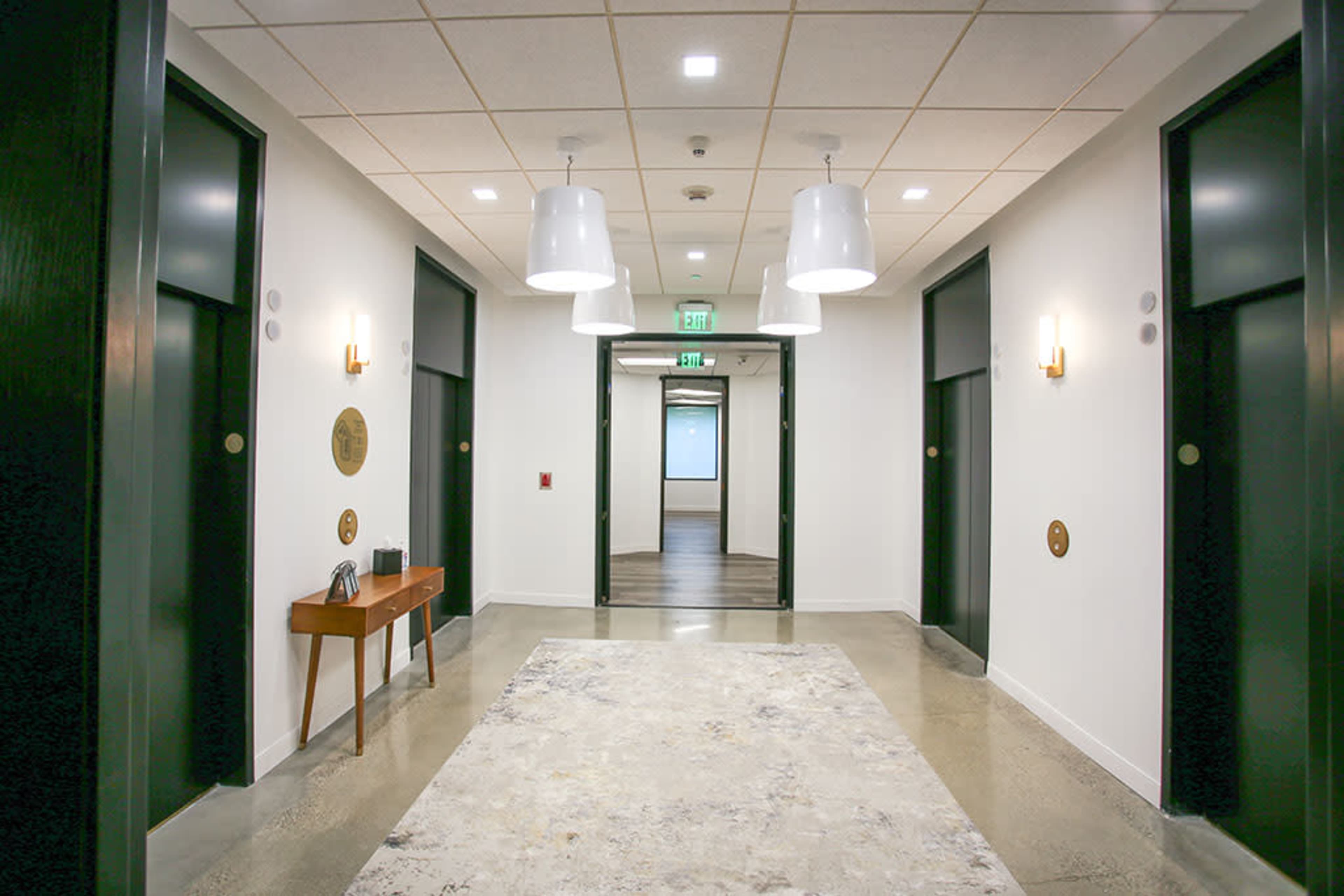 The image shows a well-lit corridor with four doorways, a wooden console table, and a large area rug on the polished concrete floor.