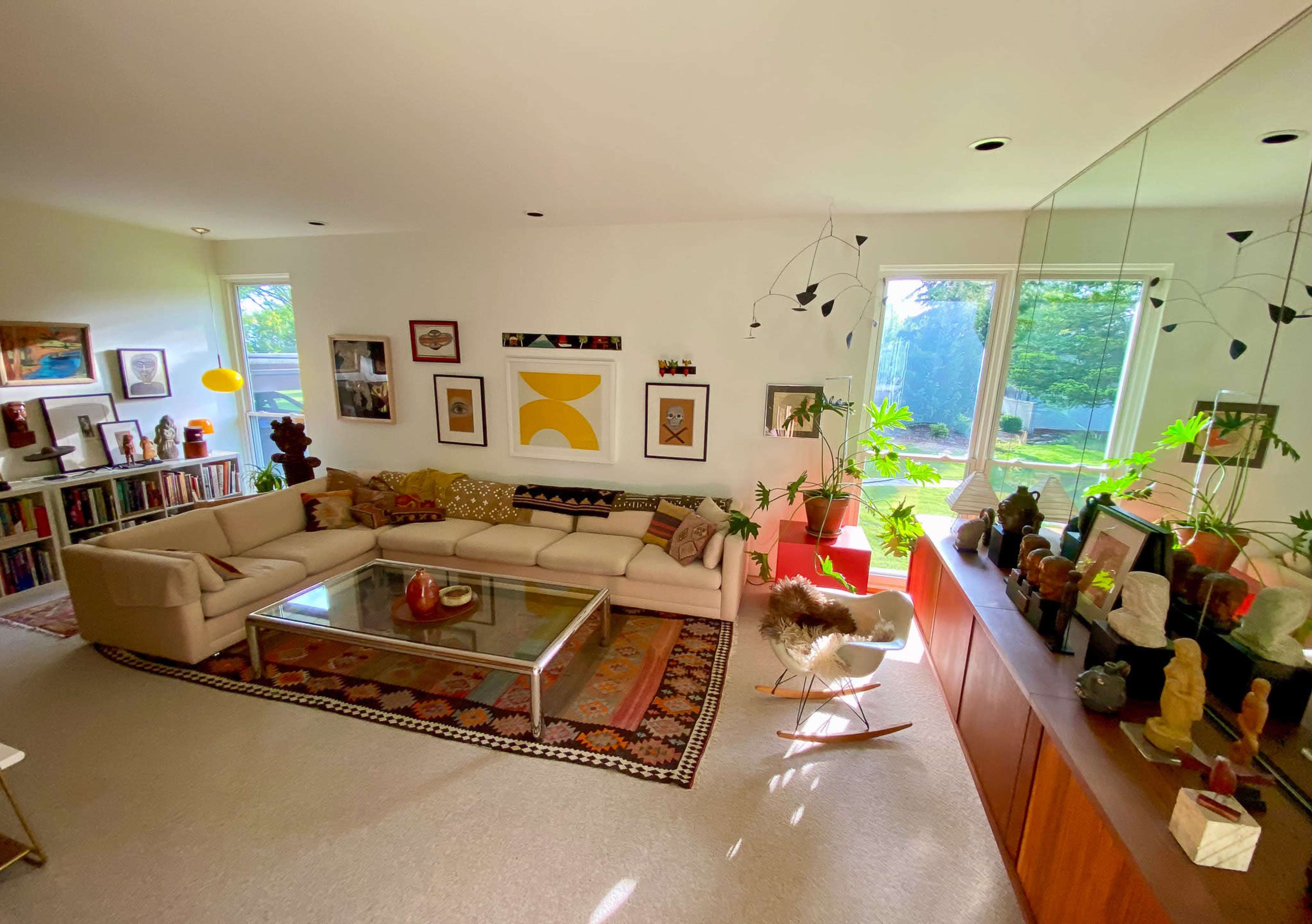 A bright living room features a large, light-colored sectional sofa, a glass coffee table, various artworks on the walls, and plants near large windows.