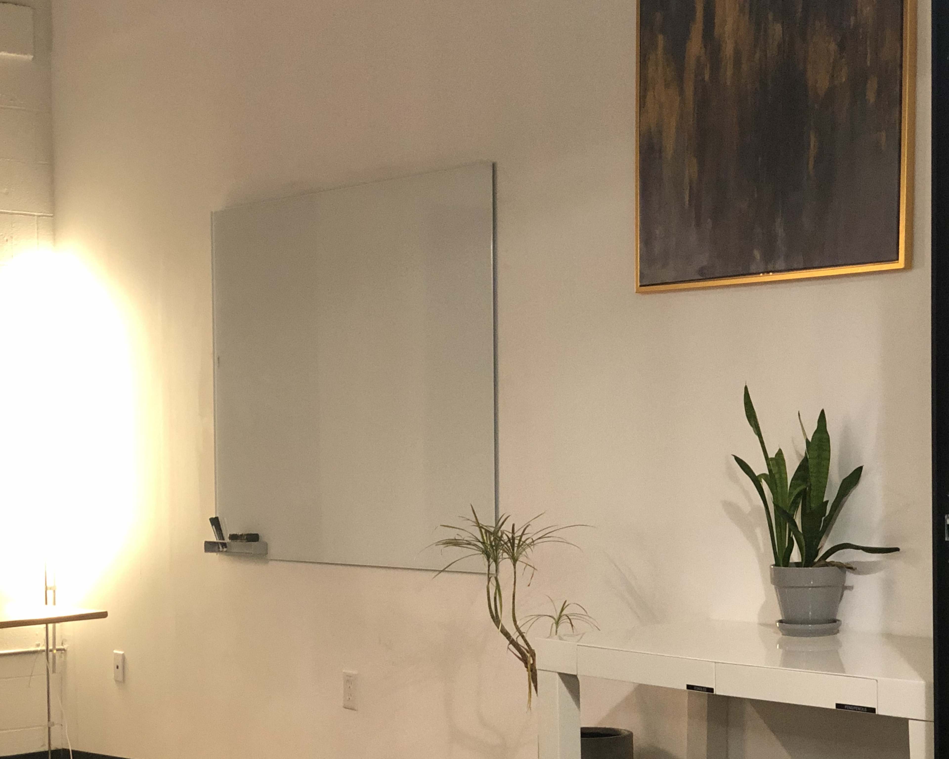 A minimalist room with a whiteboard mounted on the wall, a piece of abstract artwork above it, and a potted plant on a white table.