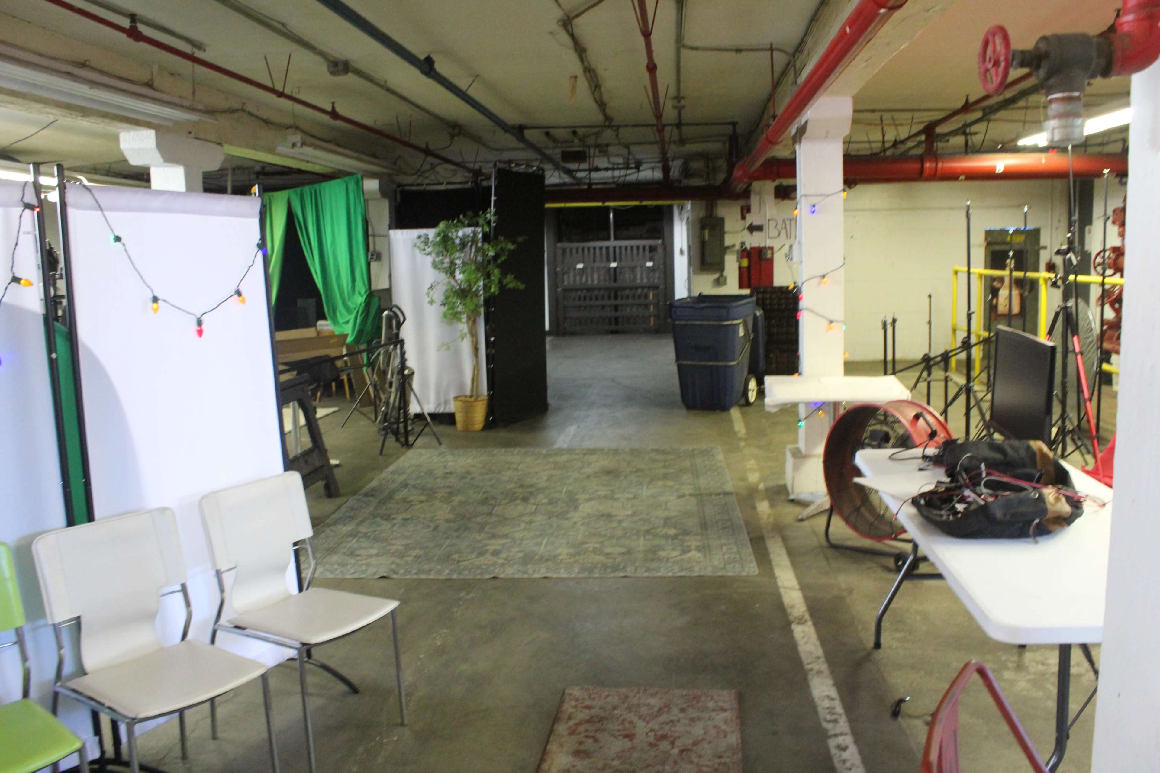 The image shows a dimly lit parking garage with various equipment and decorations, including chairs, tables, green fabric, and a potted plant.