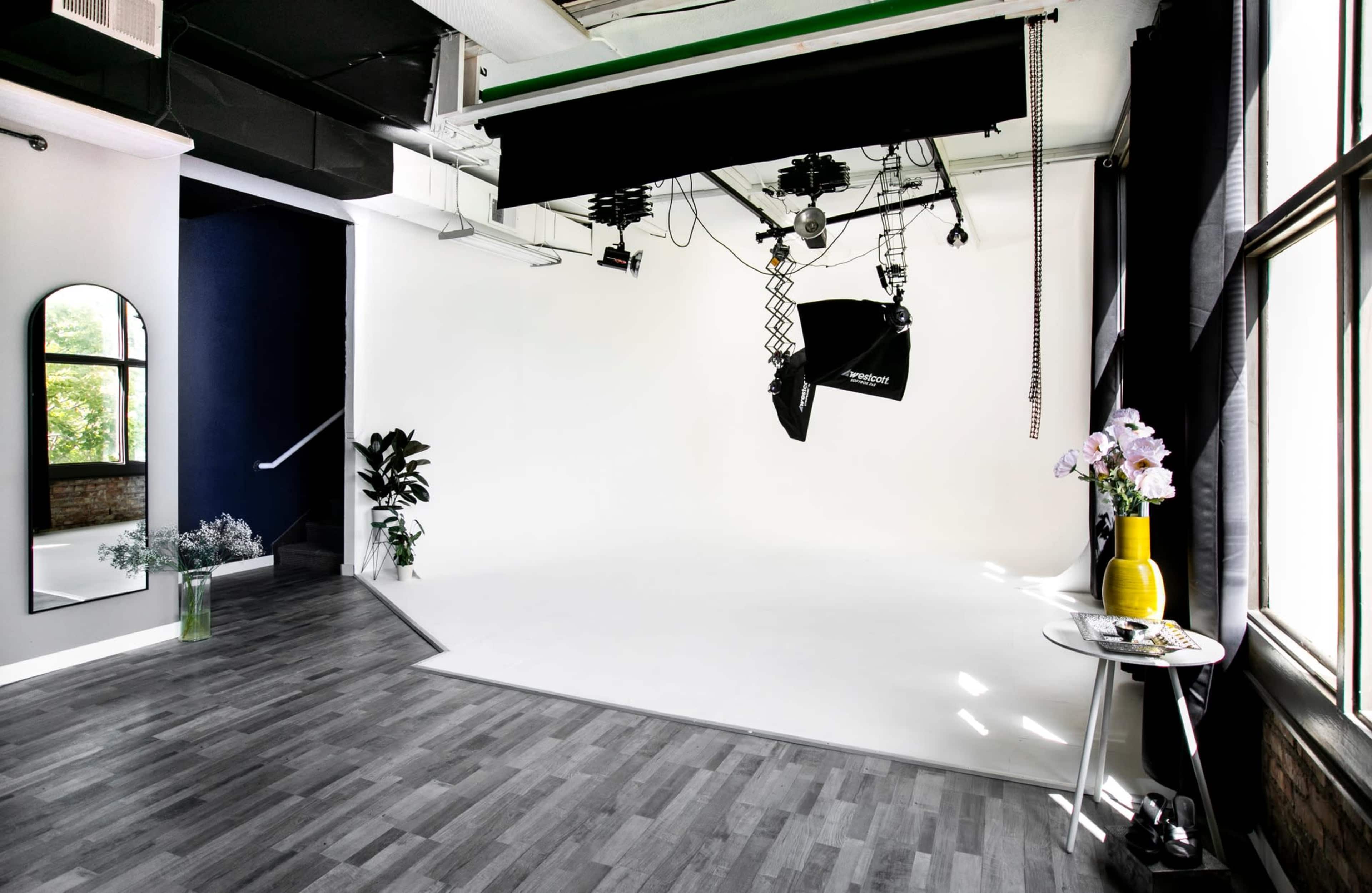 A photography studio features a white backdrop, large softbox lighting, wood flooring, and a mirror alongside decorative plants and a small table with flowers.