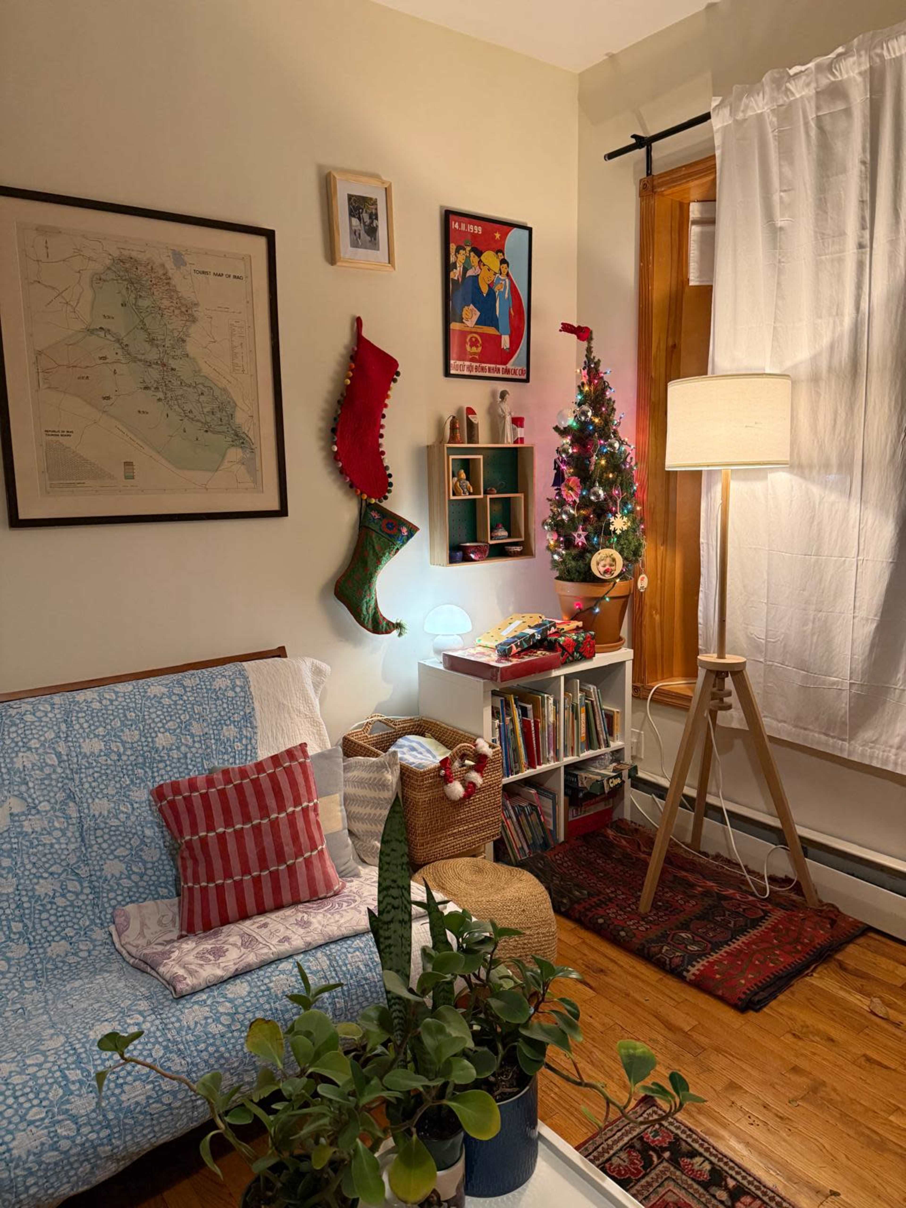 The image depicts a cozy living room corner with a small Christmas tree, decorative stockings, and a bookshelf next to a lamp on a wooden stand.