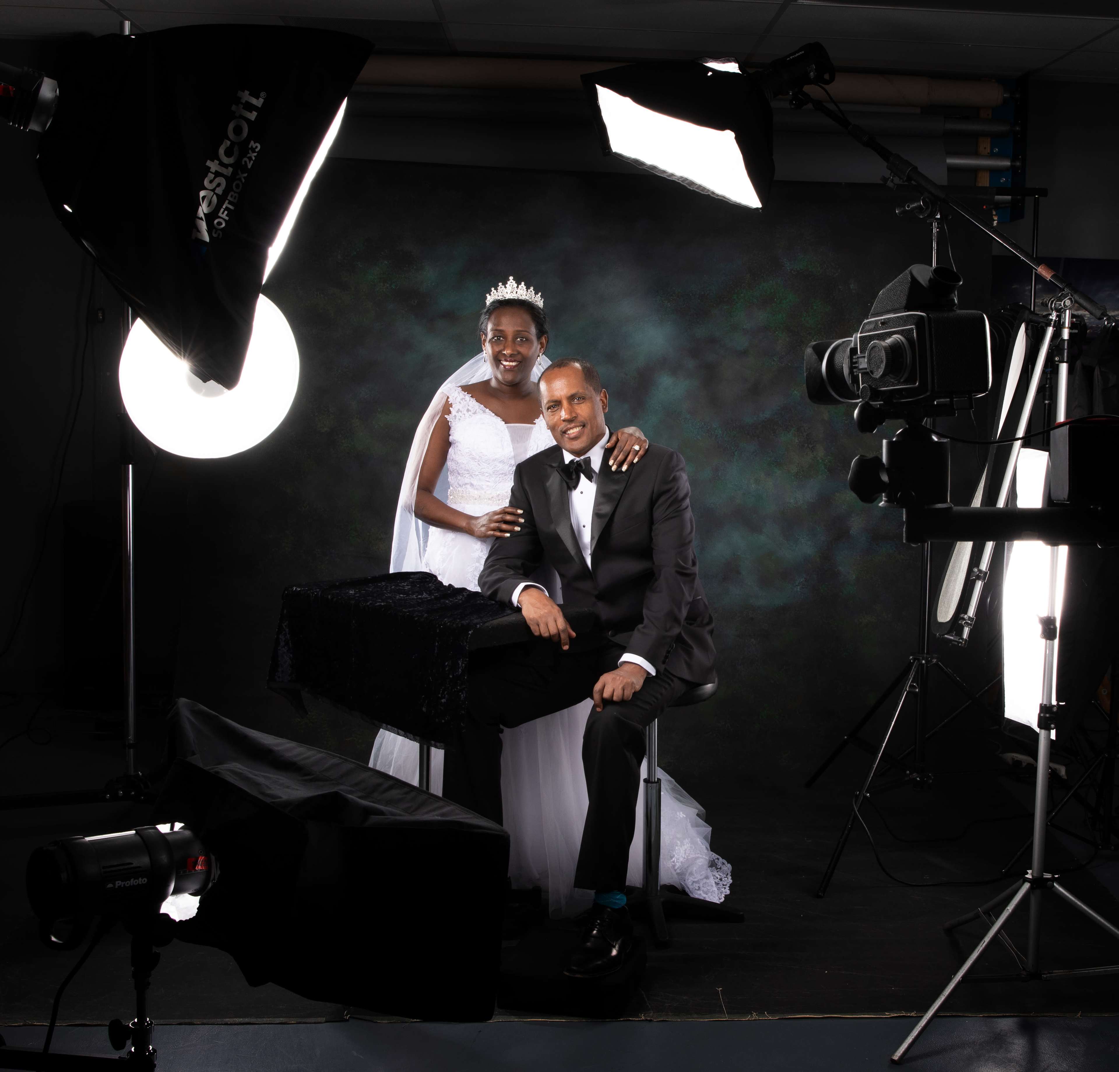 A bride in a white gown stands behind a groom in a tuxedo, both posing for a formal portrait in a professional photography studio surrounded by various lighting equipment.