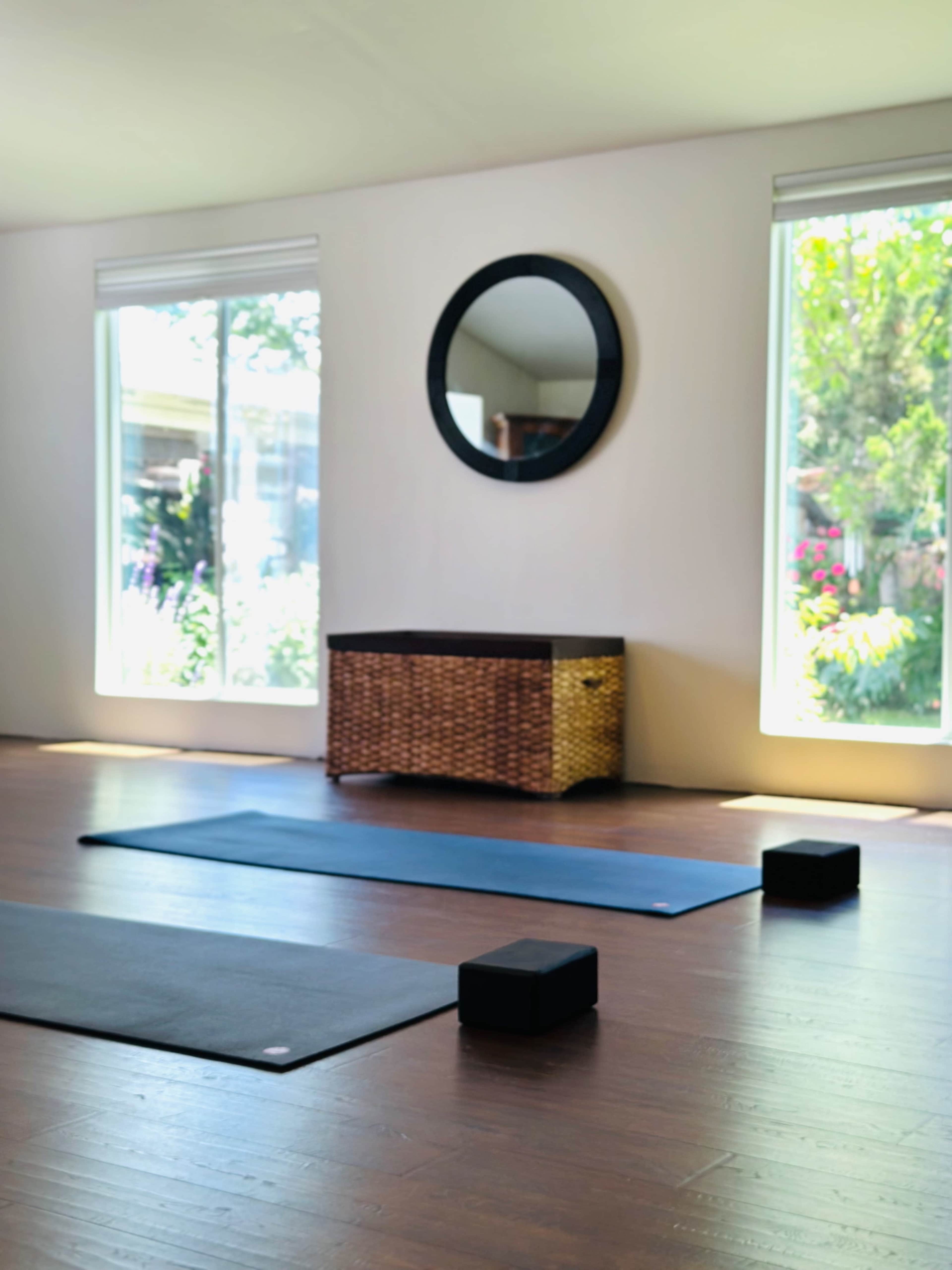 The image shows a yoga studio with two mats, two yoga blocks, a round mirror, and a wicker storage box, illuminated by natural light from large windows.