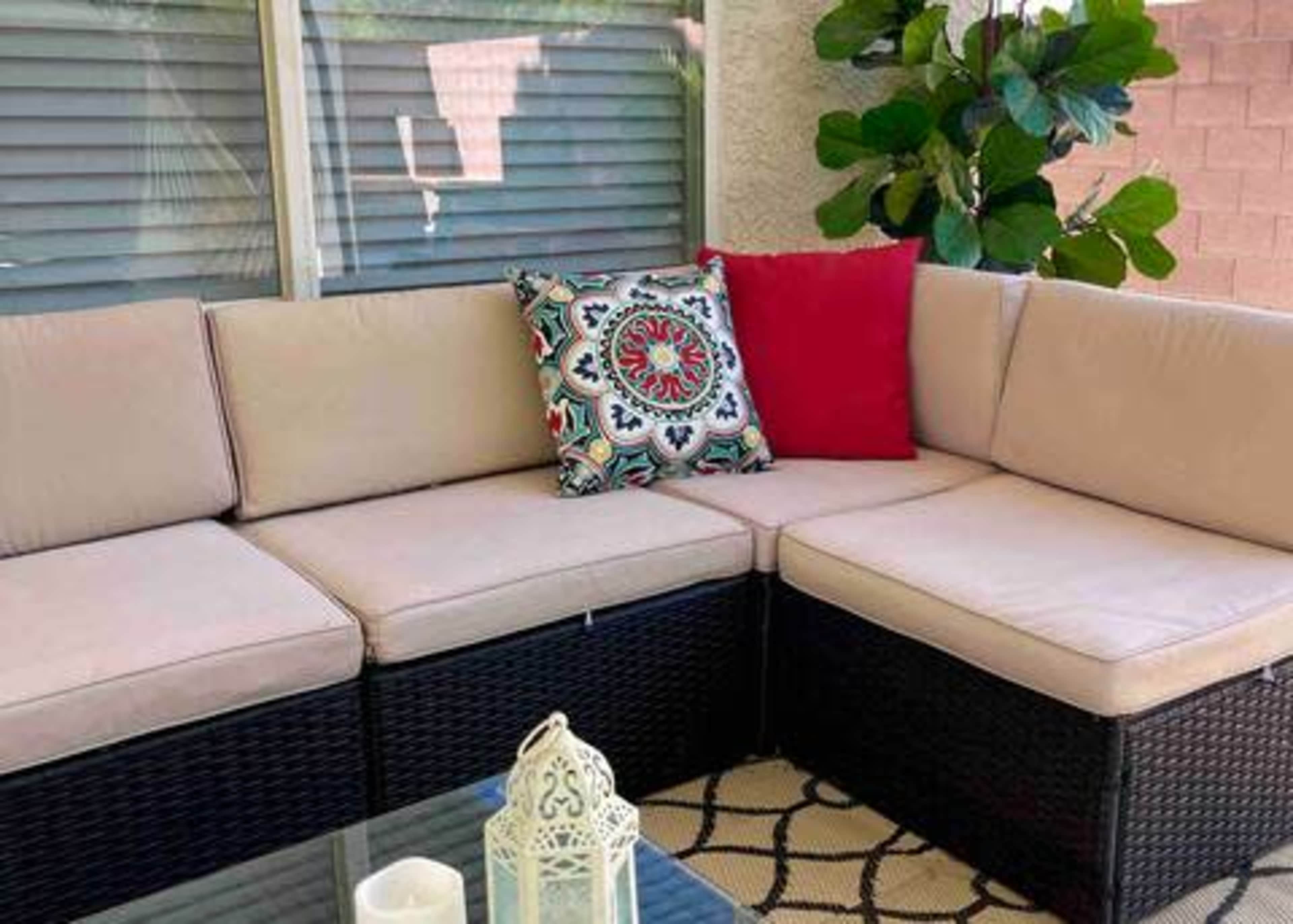 A modern outdoor seating area features a sectional couch with beige cushions, a decorative pillow, and a glass-top table, surrounded by greenery.