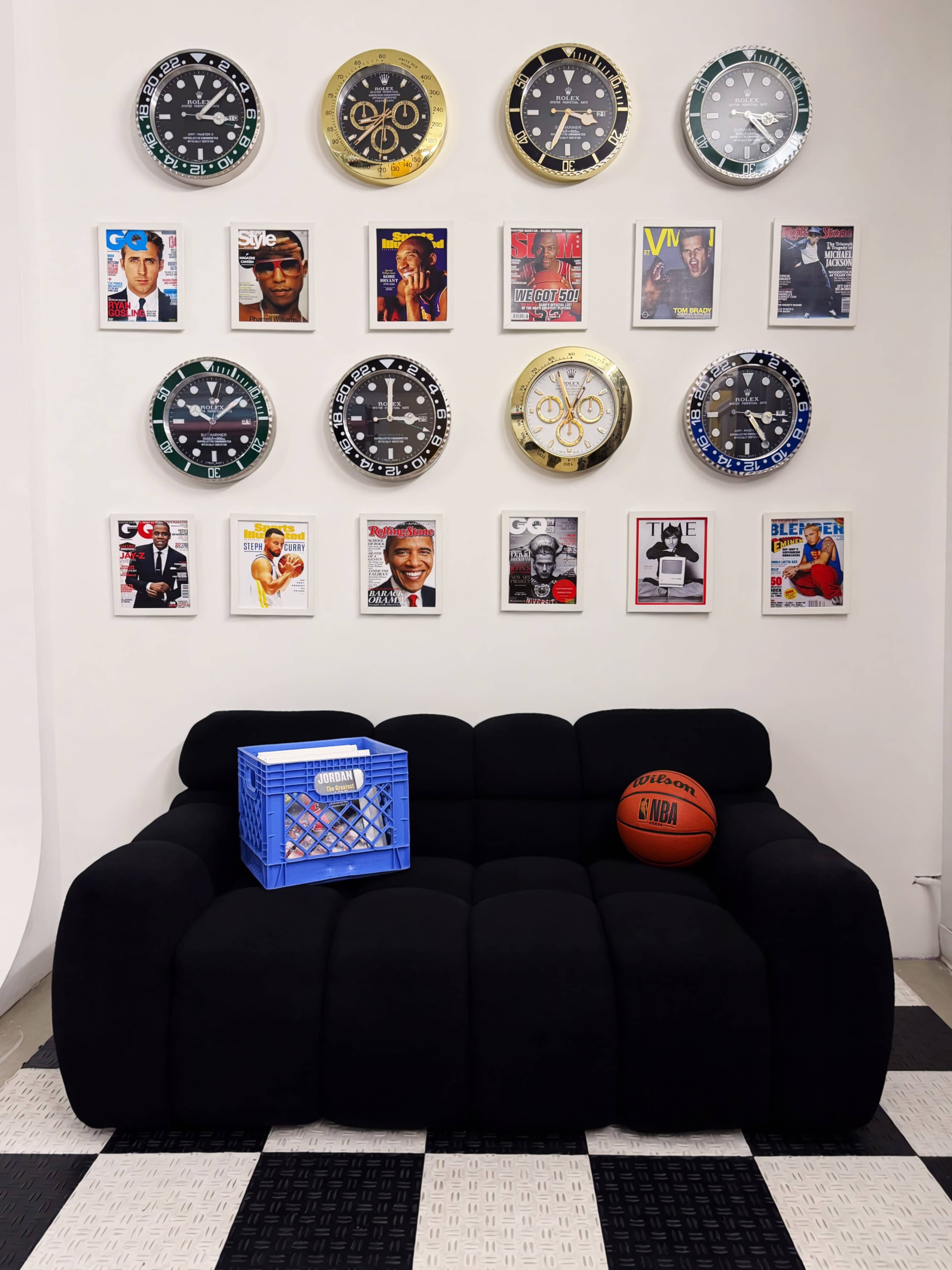 The image features a black couch in front of a wall adorned with various wall clocks and magazine covers, with a blue crate and a basketball placed on the couch.