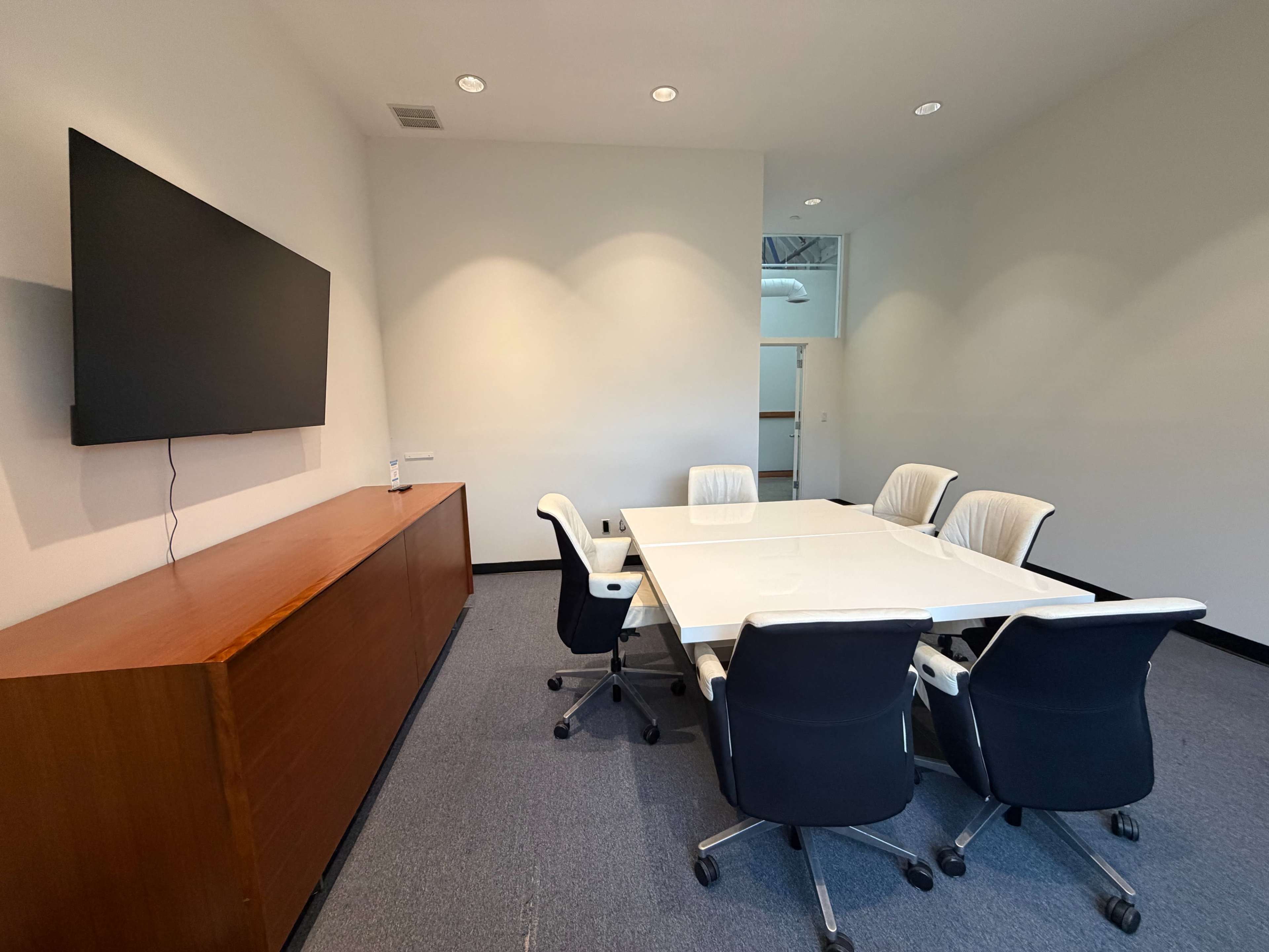The image shows a modern conference room featuring a large white table surrounded by ergonomic chairs, with a television mounted on the wall and a wooden sideboard.