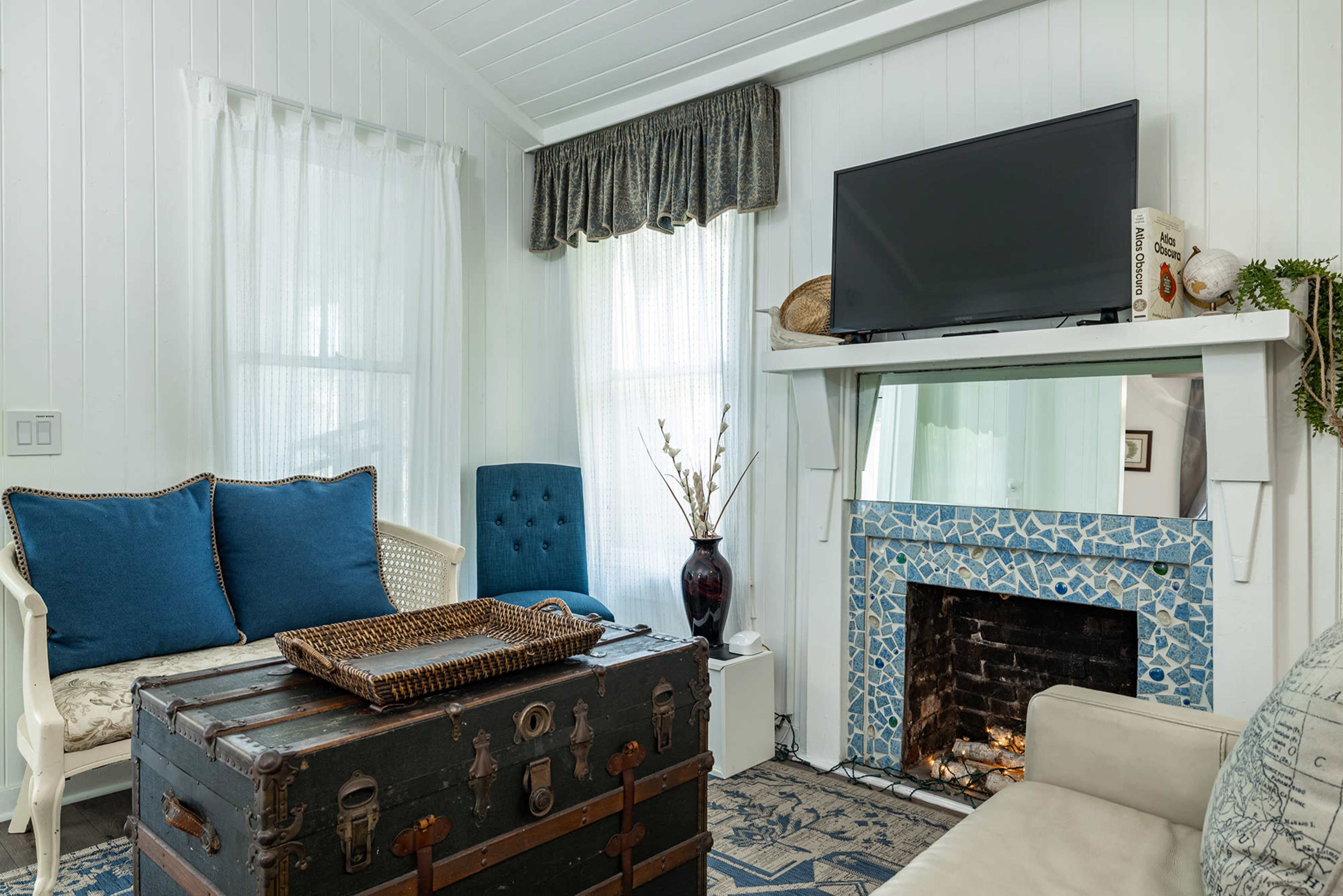The image shows a cozy living room with a blue and white color scheme, featuring a vintage trunk, a television mounted above a mosaic fireplace, and a mix of seating options.
