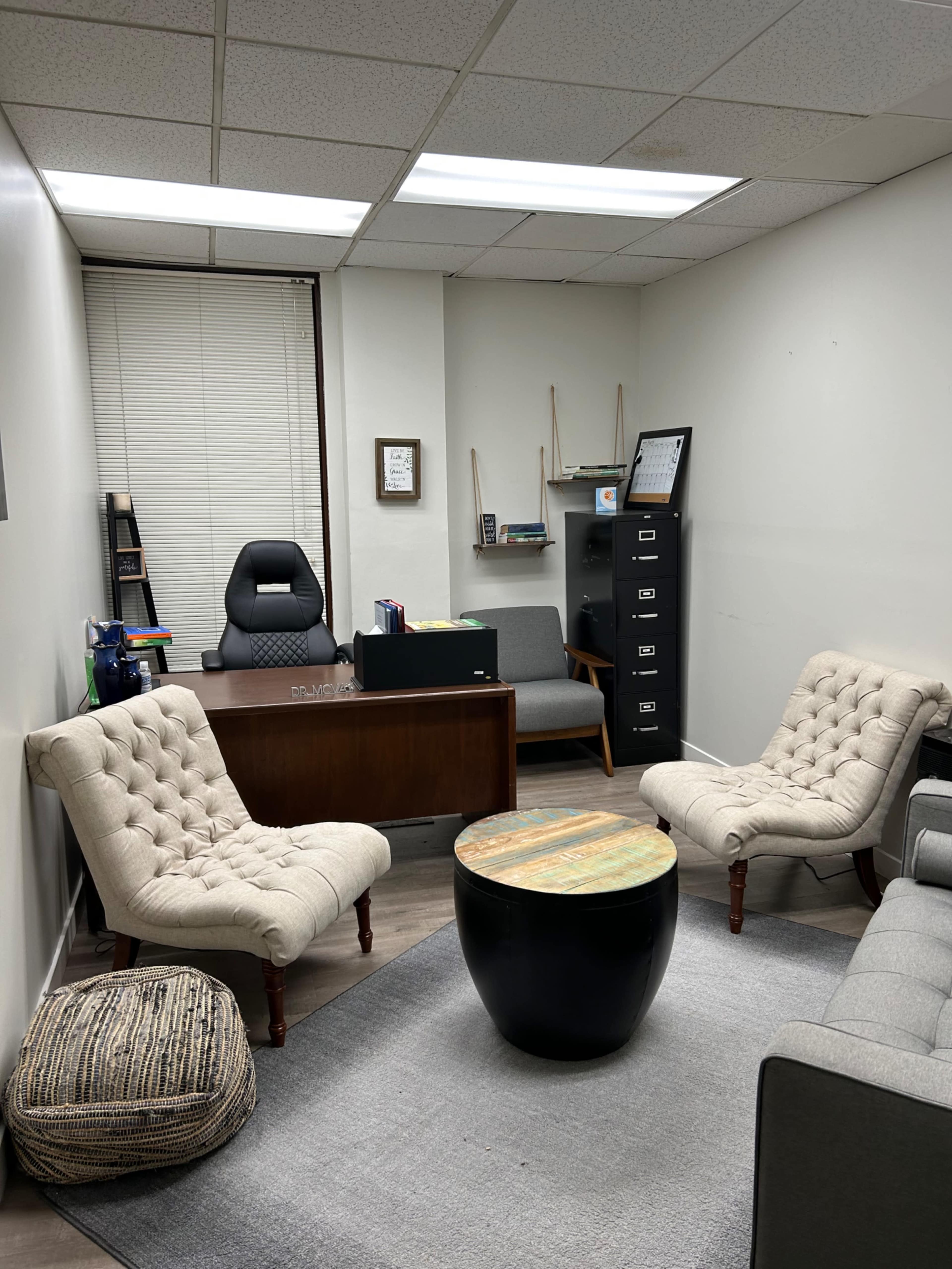 The image shows an office space with two upholstered chairs, a round coffee table, a desk with a chair, and shelving containing various items.