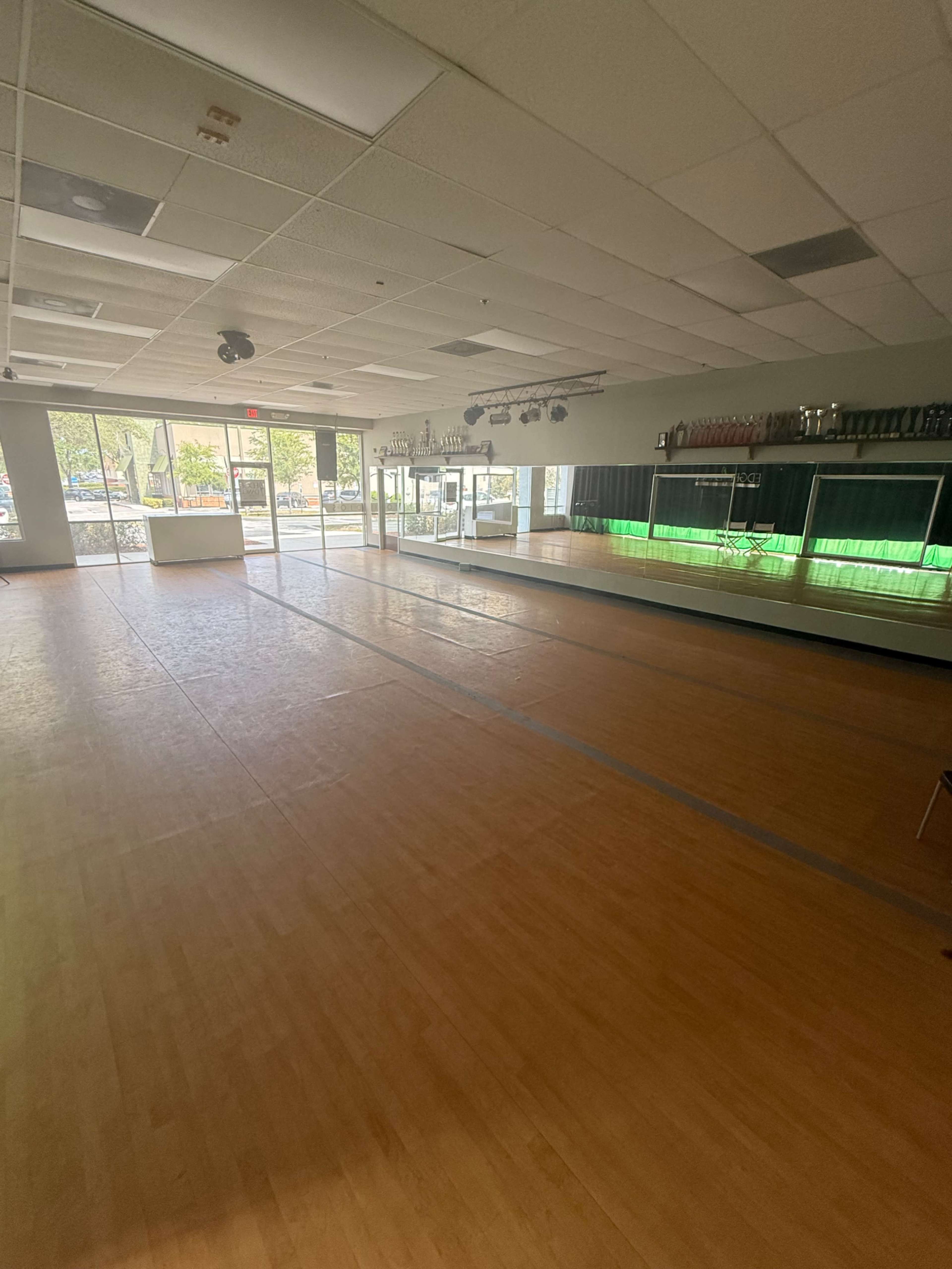 The image shows a spacious dance studio with wooden flooring, large mirrors, and bright windows allowing natural light to enter.