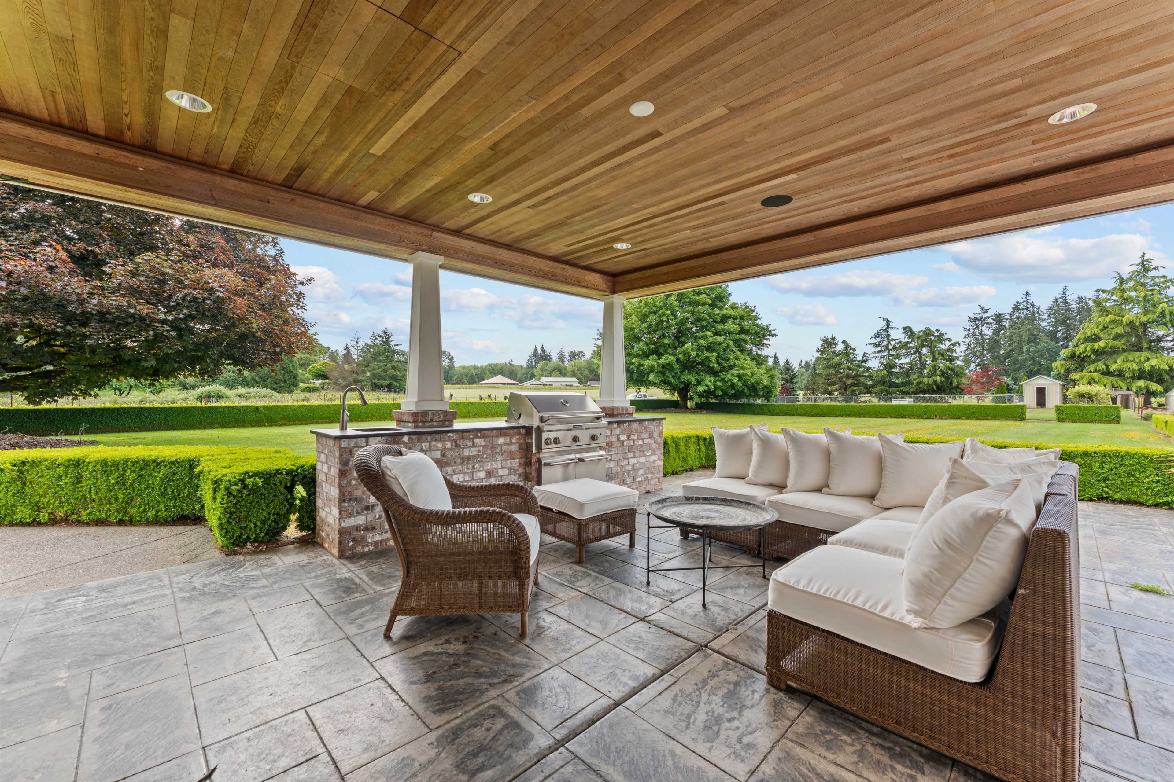 The image shows a covered outdoor seating area with a wicker sofa and gray coffee table, overlooking a garden with neatly trimmed hedges.