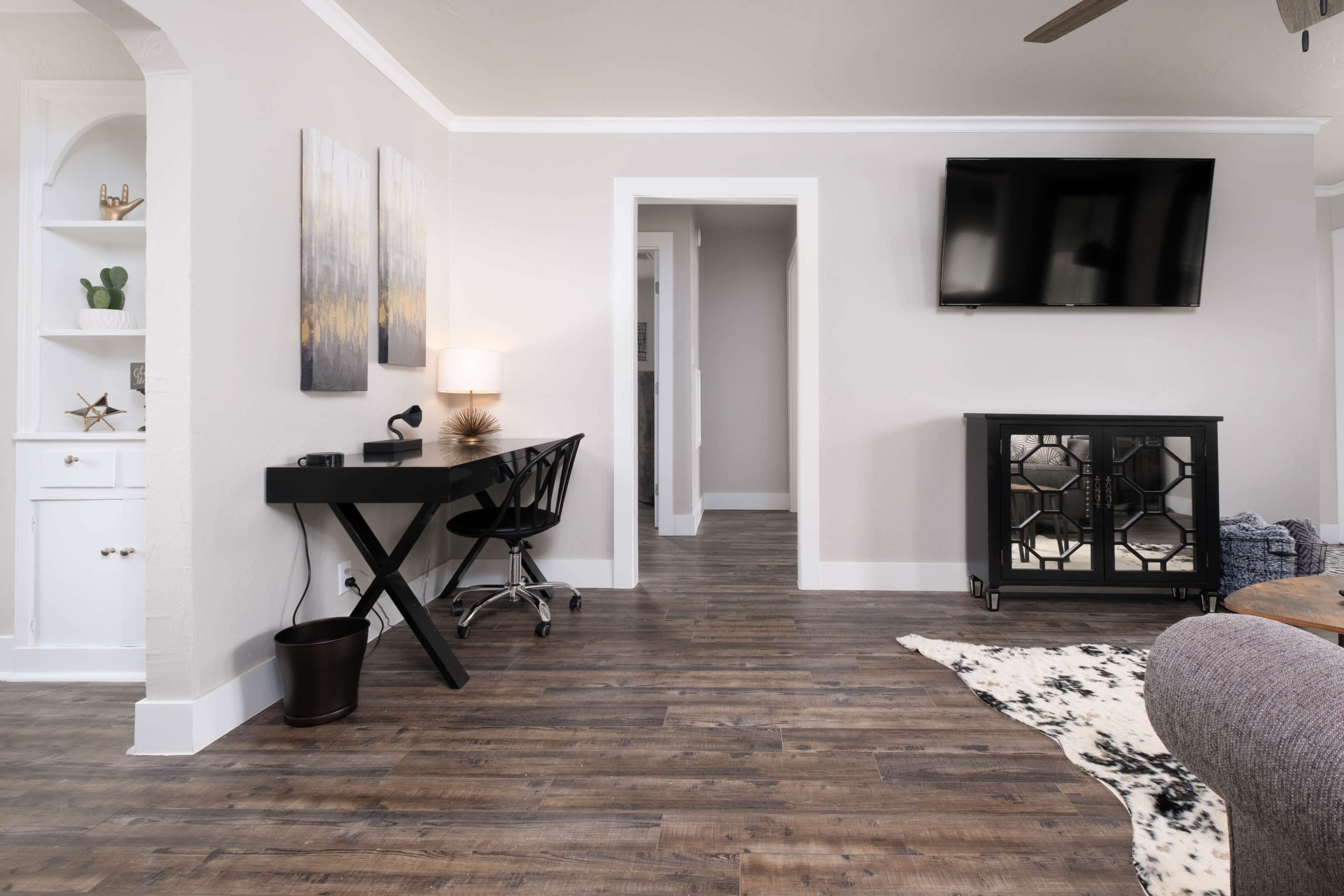The image shows a modern living space with a black desk and chair, a television mounted on the wall, and a hallway leading to another room.