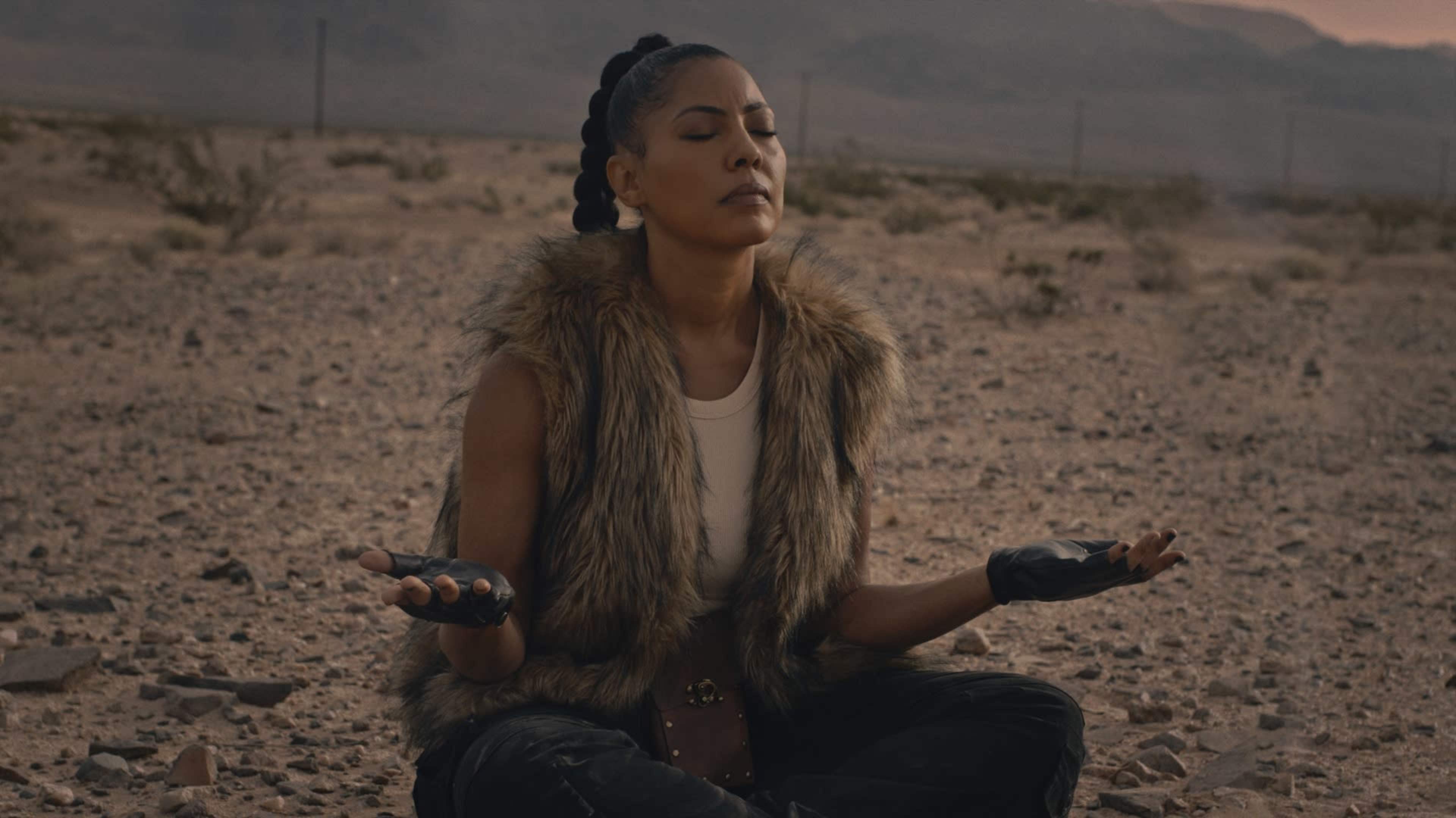 A woman sits cross-legged on a rocky desert floor, with her eyes closed and hands resting on her knees.