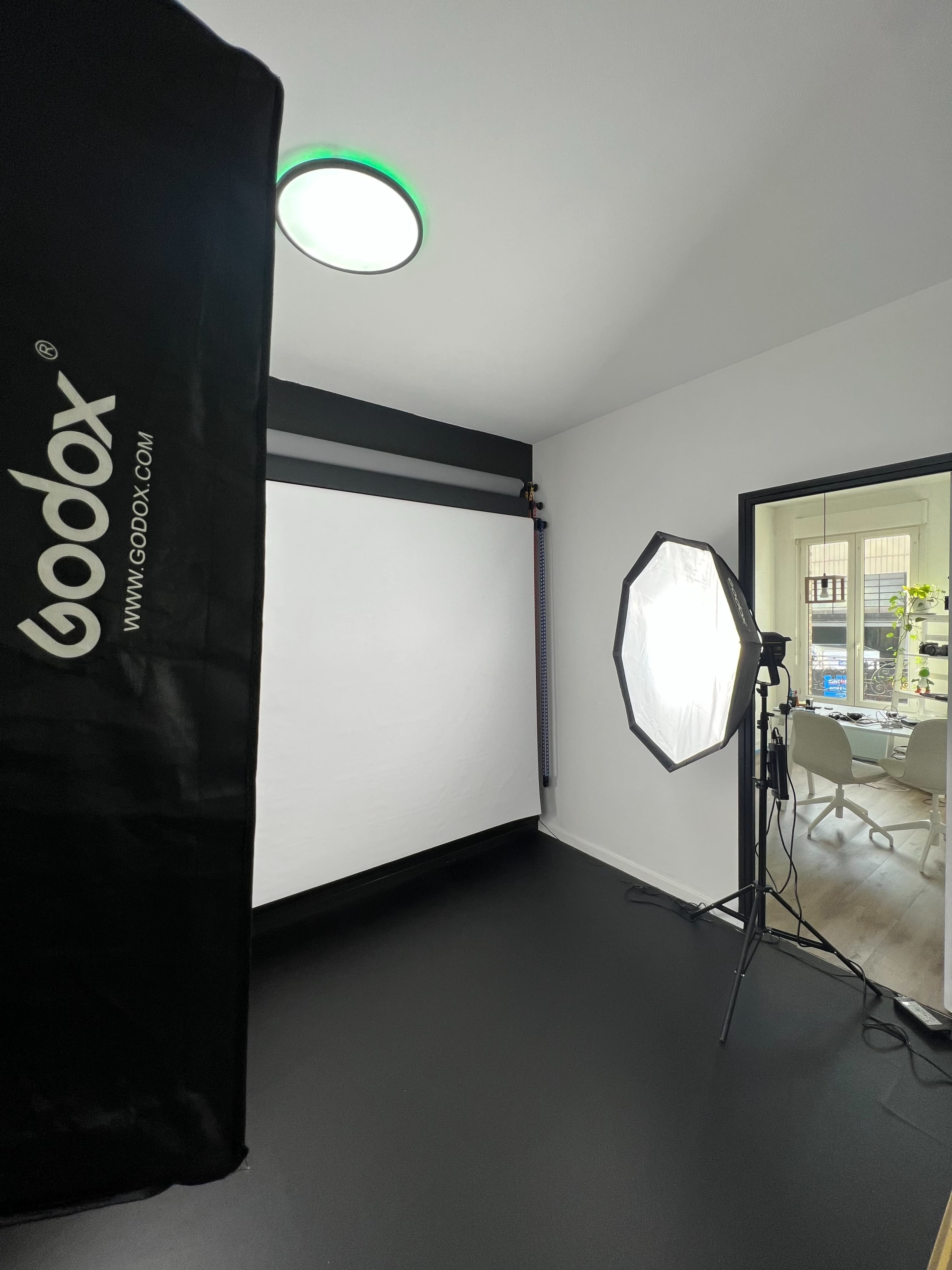 A photography studio setup featuring a softbox light, a white backdrop, and a doorway leading to a room with visible furniture.