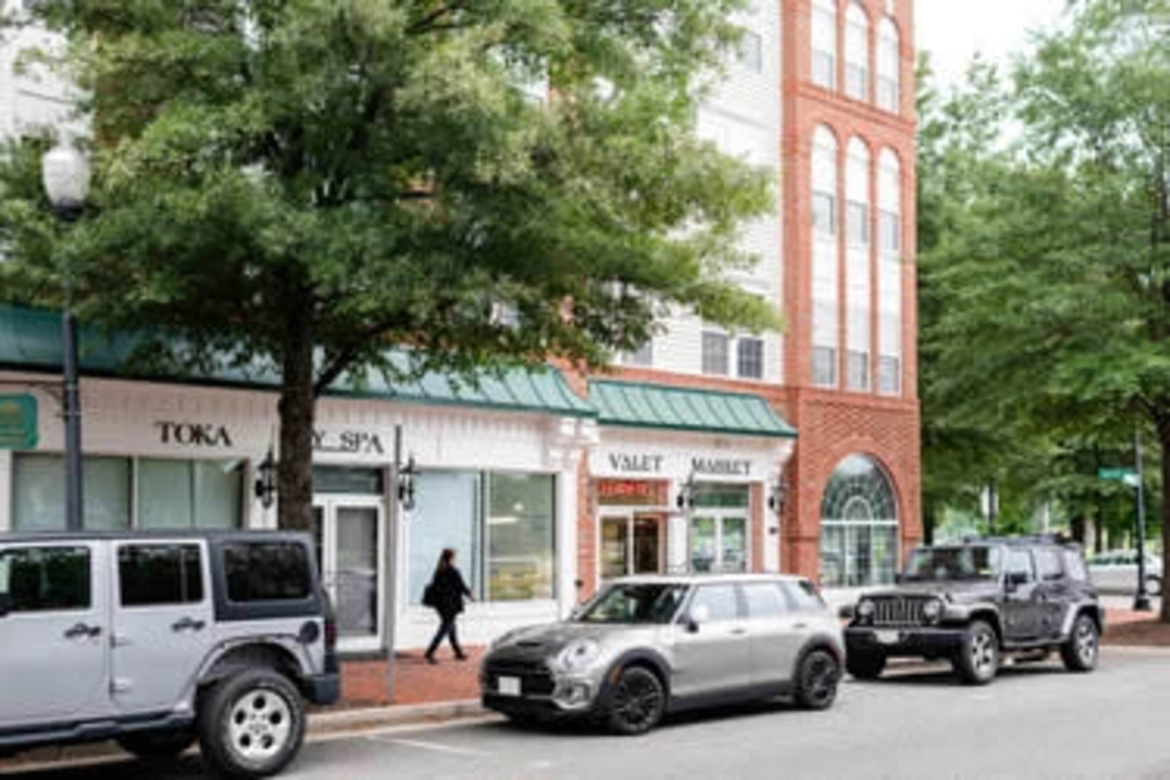 A street scene with parked vehicles, including a black Mini Cooper and several SUVs, in front of shops with large windows and greenery lining the sidewalk.