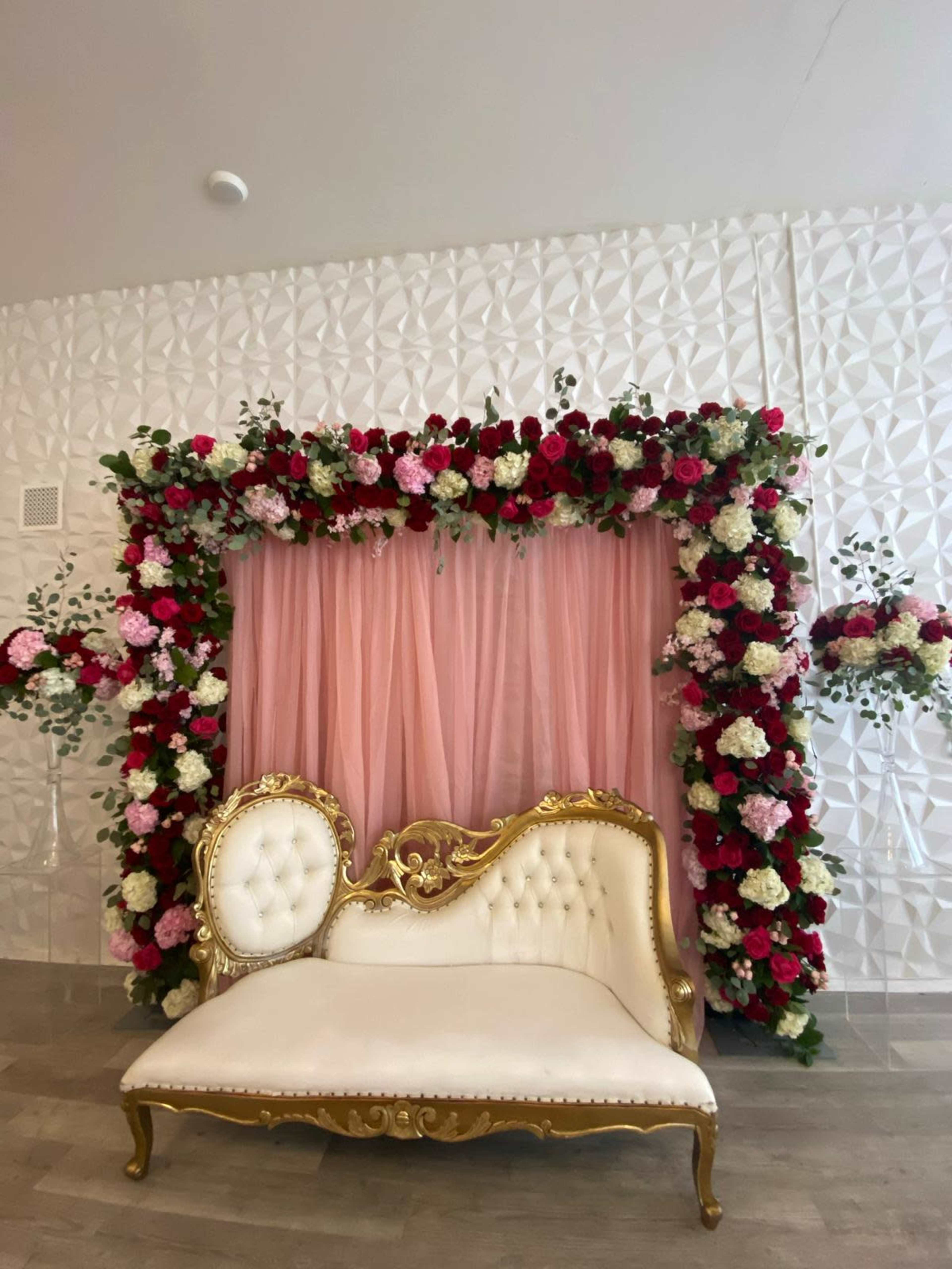 A decorative backdrop of pink fabric adorned with an arrangement of red, white, and pink flowers, accompanied by an ornate white and gold sofa in front.