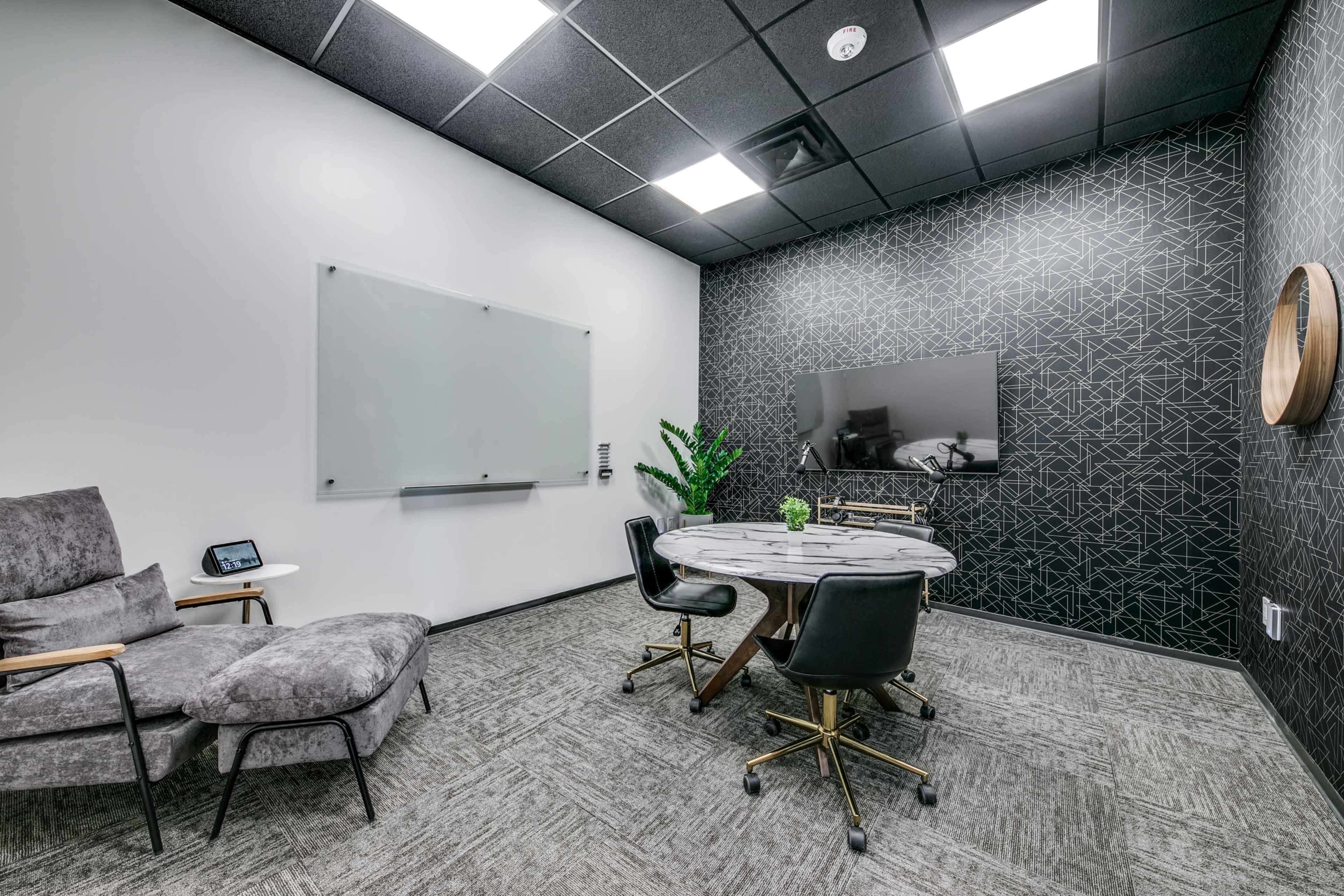 A small meeting room featuring a round table surrounded by four black chairs, a gray lounge chair, a whiteboard, and a TV on a patterned wall.