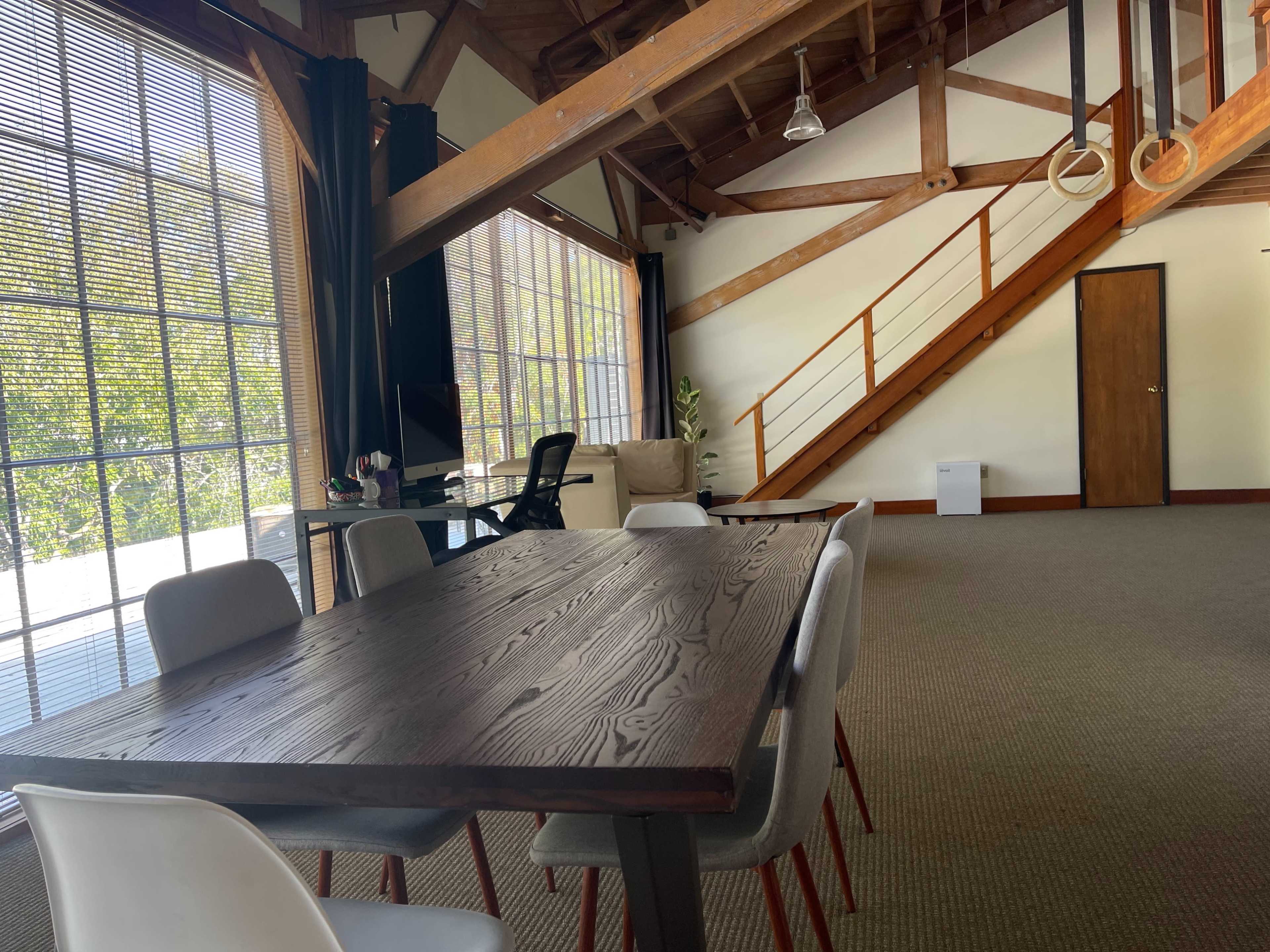 A spacious interior with a wooden table surrounded by chairs, large windows letting in natural light, and an upper staircase leading to a loft area.