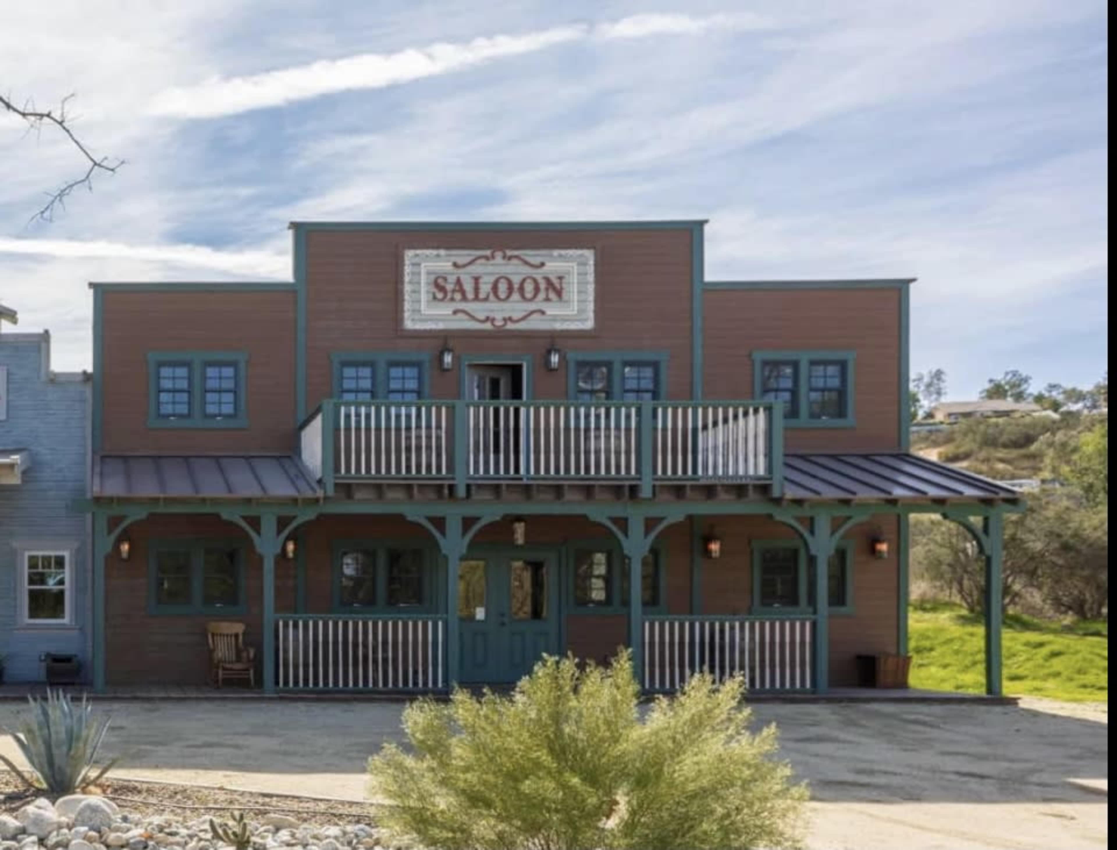 A two-story wooden saloon with a covered balcony and large windows, set in a rural landscape.