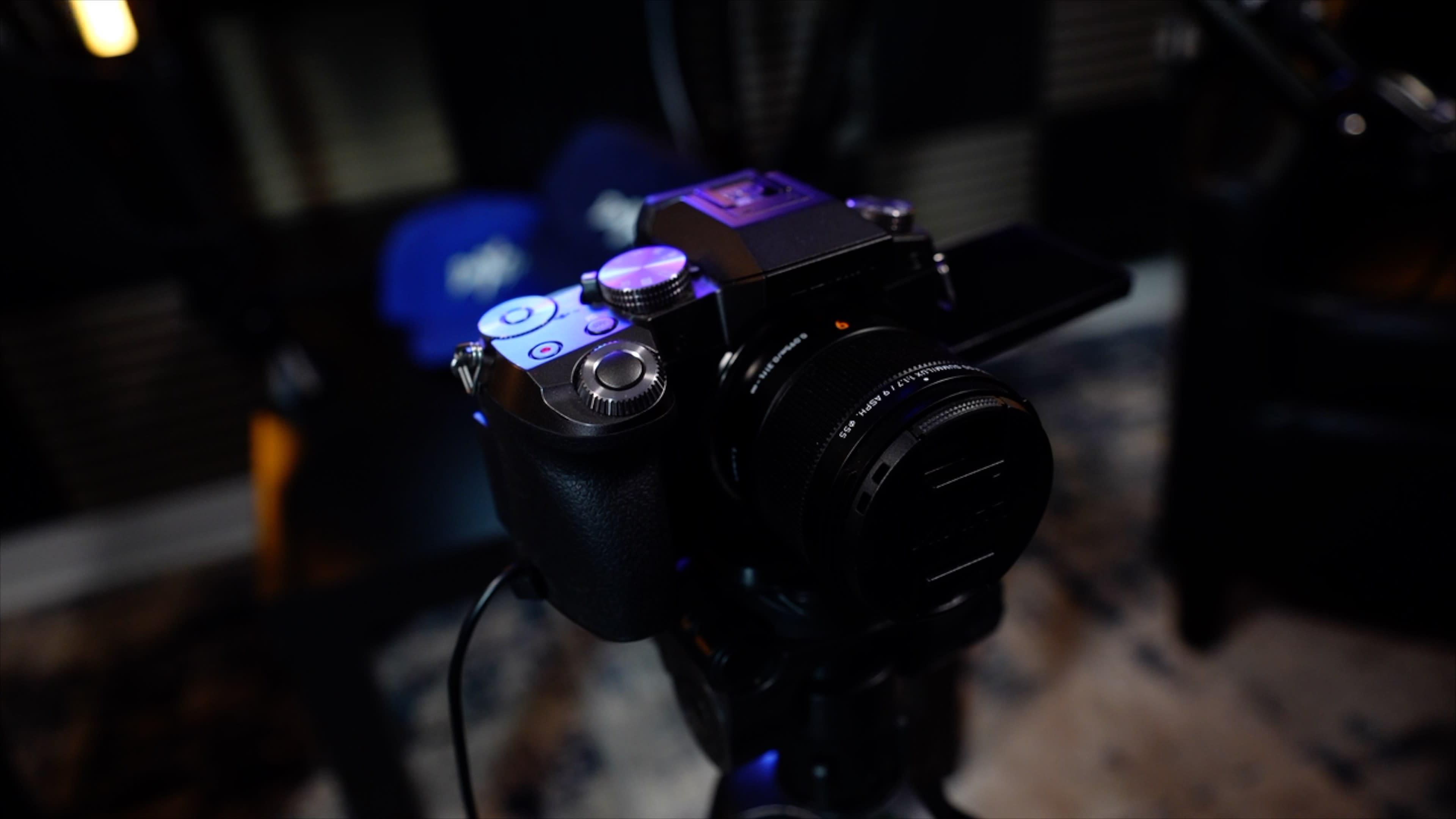 A camera is mounted on a tripod with a blurred background of hats and studio equipment.