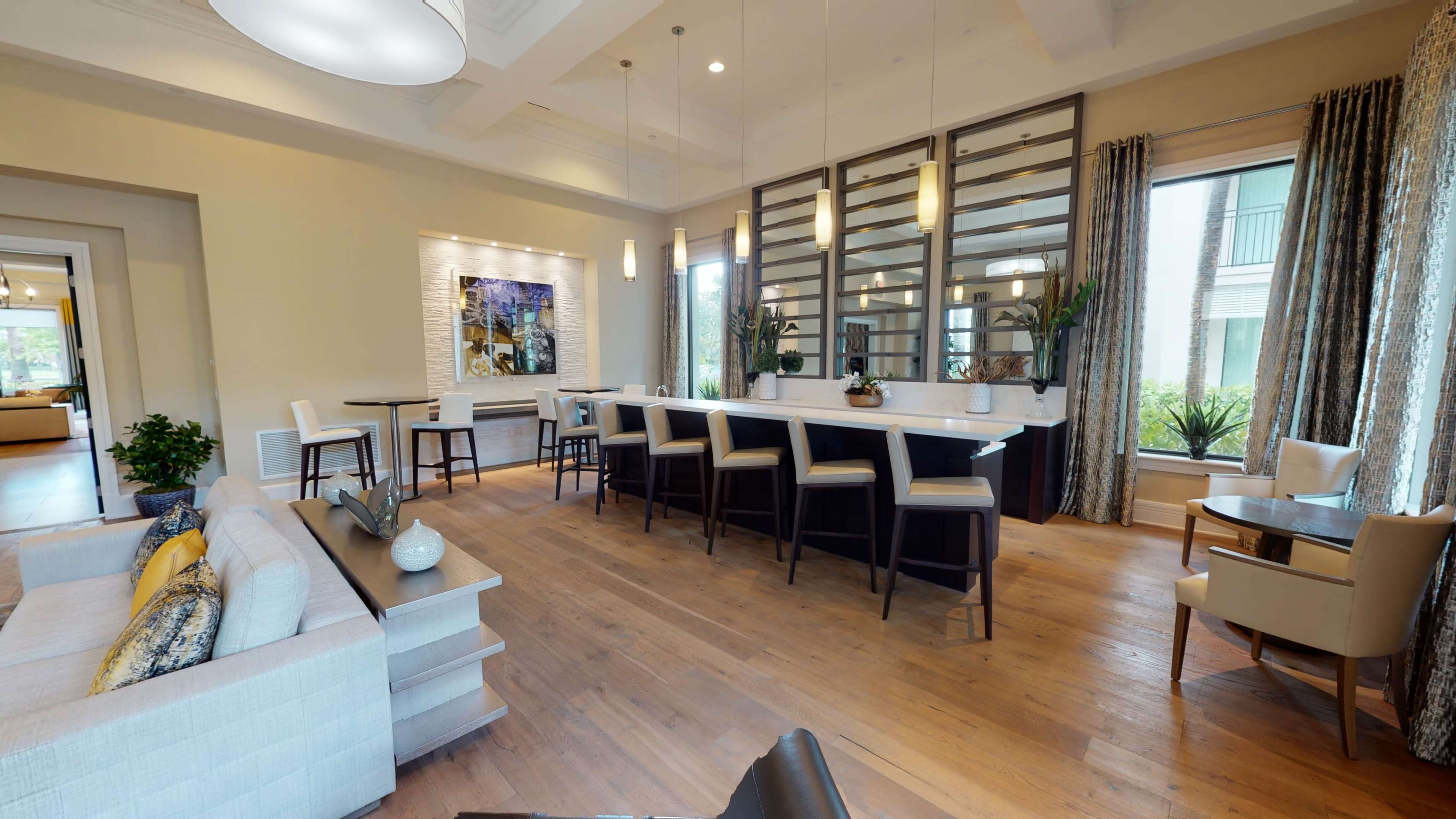 The image shows a spacious, well-lit room featuring a seating area, a bar-style counter with stools, and large windows with plants outside.