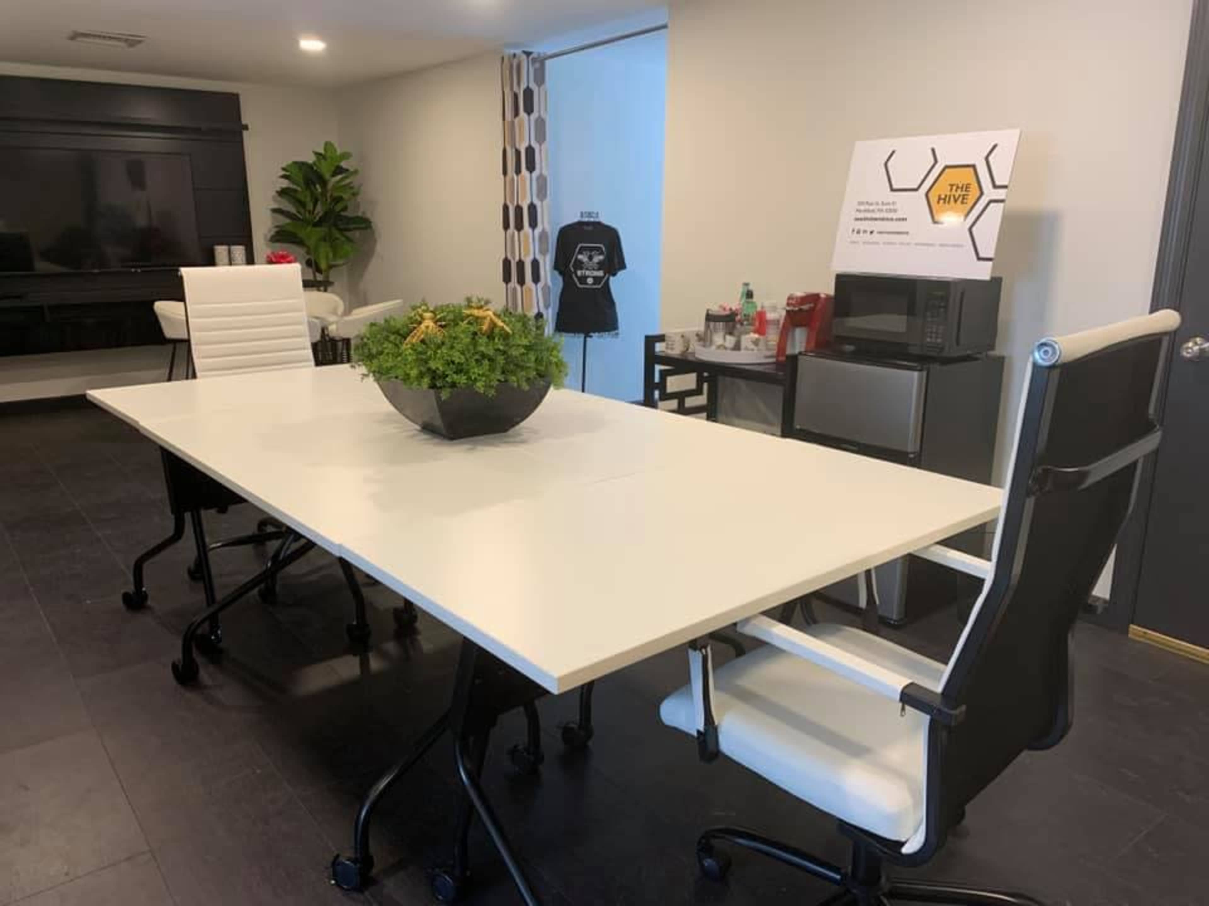 A modern conference room features a large white table surrounded by black and white chairs, with a potted plant centerpiece and a sign displaying "THE HIVE."