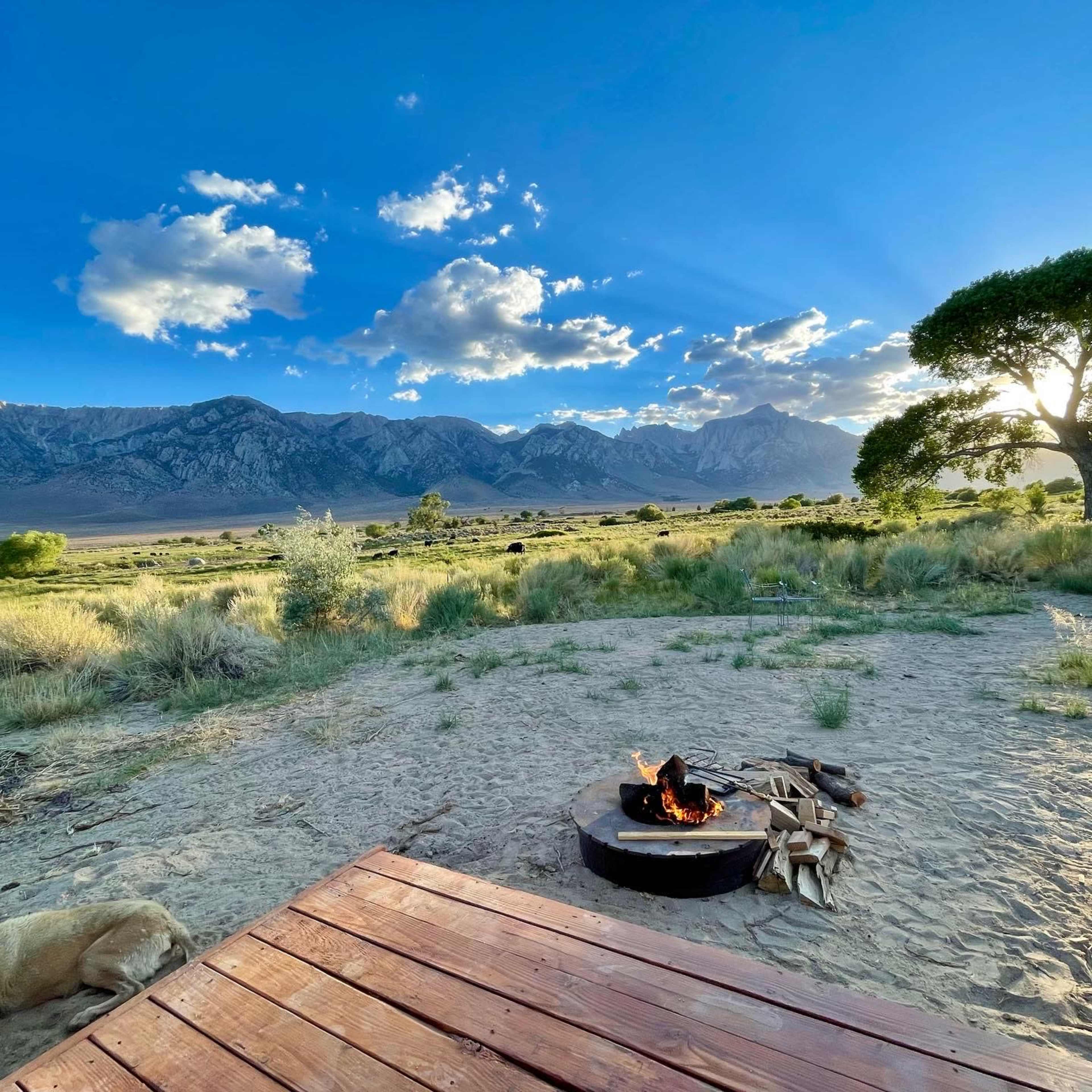 A campfire burns in a circular pit surrounded by wooden decking, with mountains and a blue sky in the background.