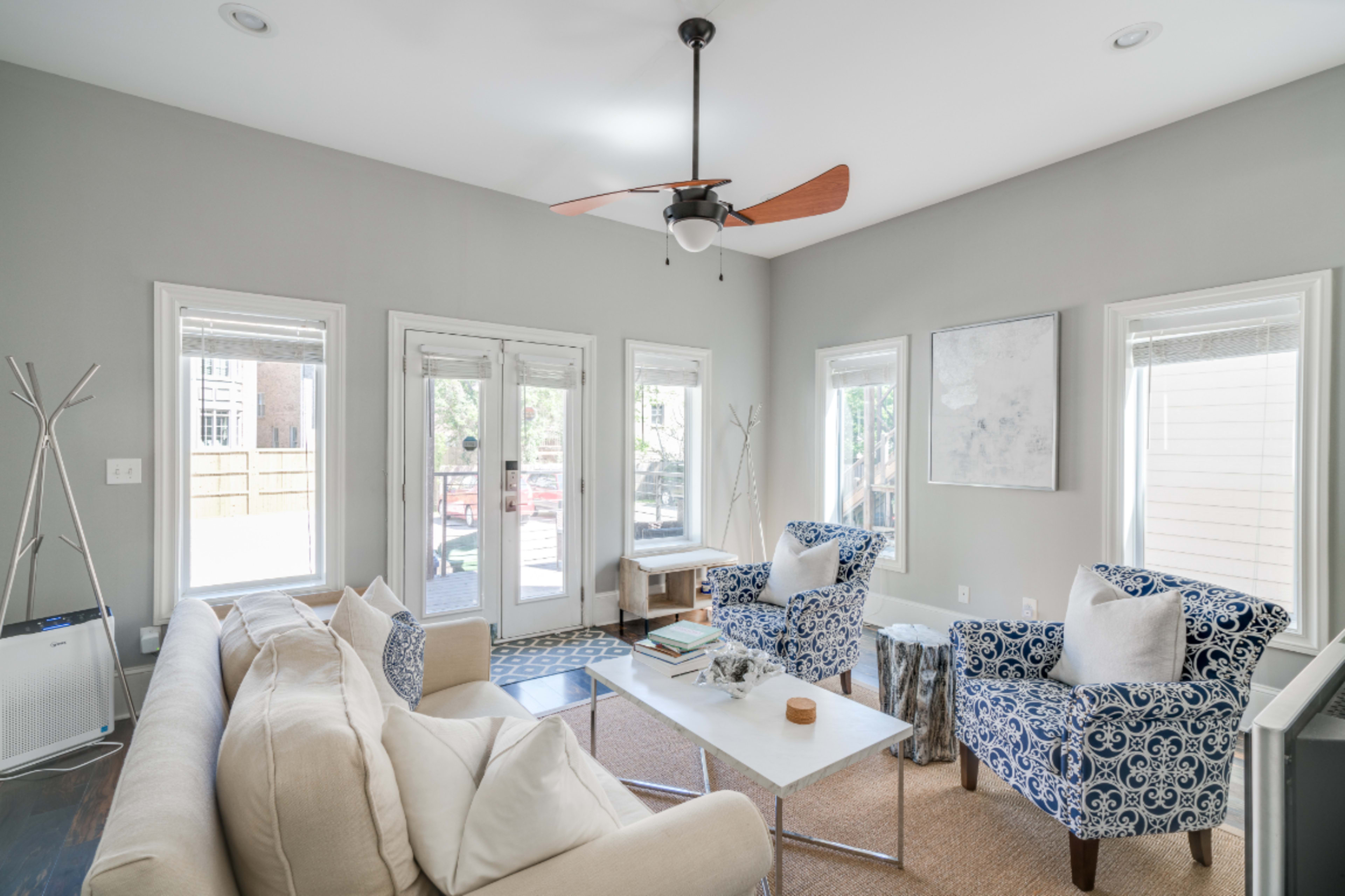 The image shows a modern living room with a beige sofa, blue patterned armchairs, a coffee table, and large windows allowing natural light.