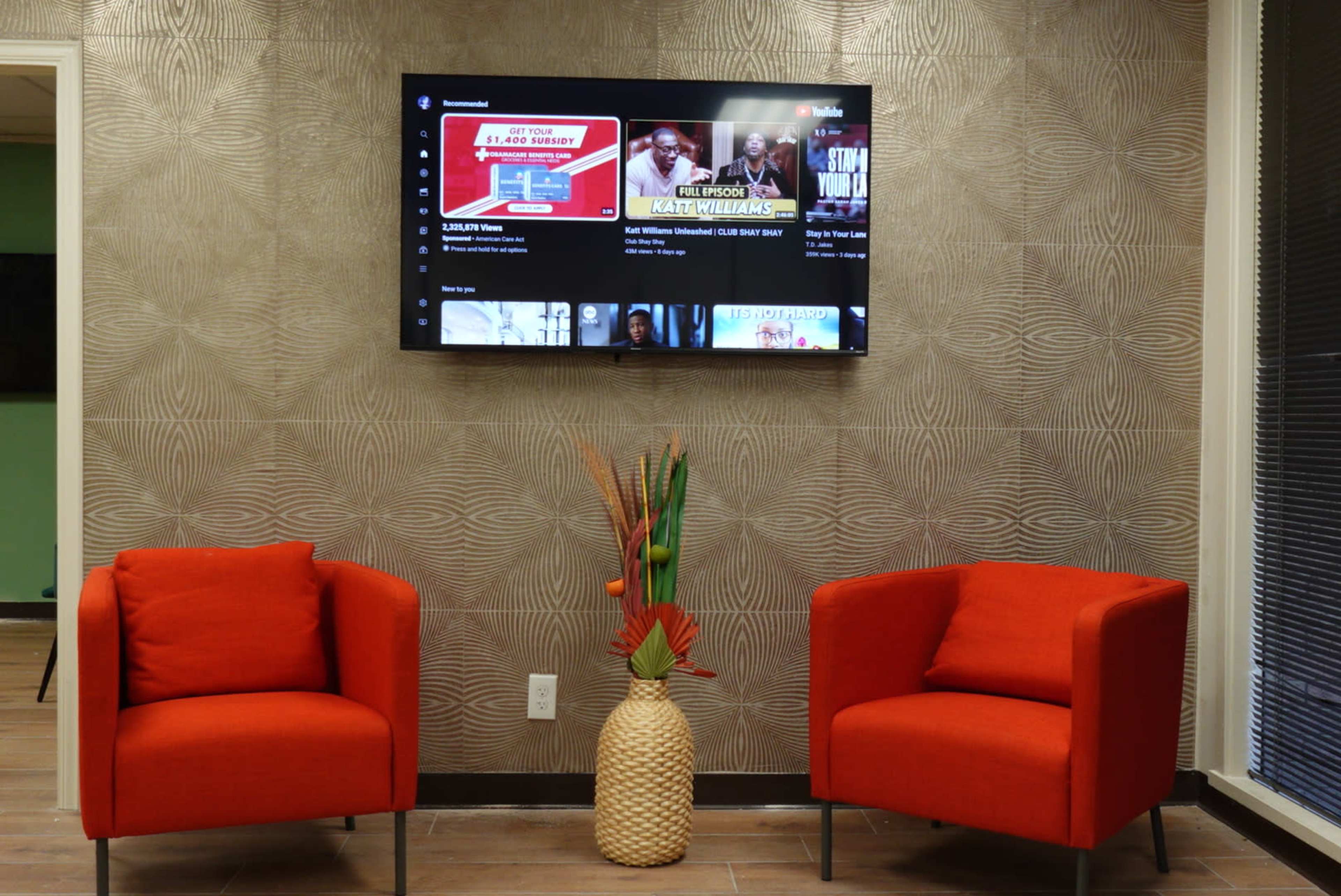 Two orange chairs positioned in front of a textured wall, with a television mounted above displaying a selection of video thumbnails.