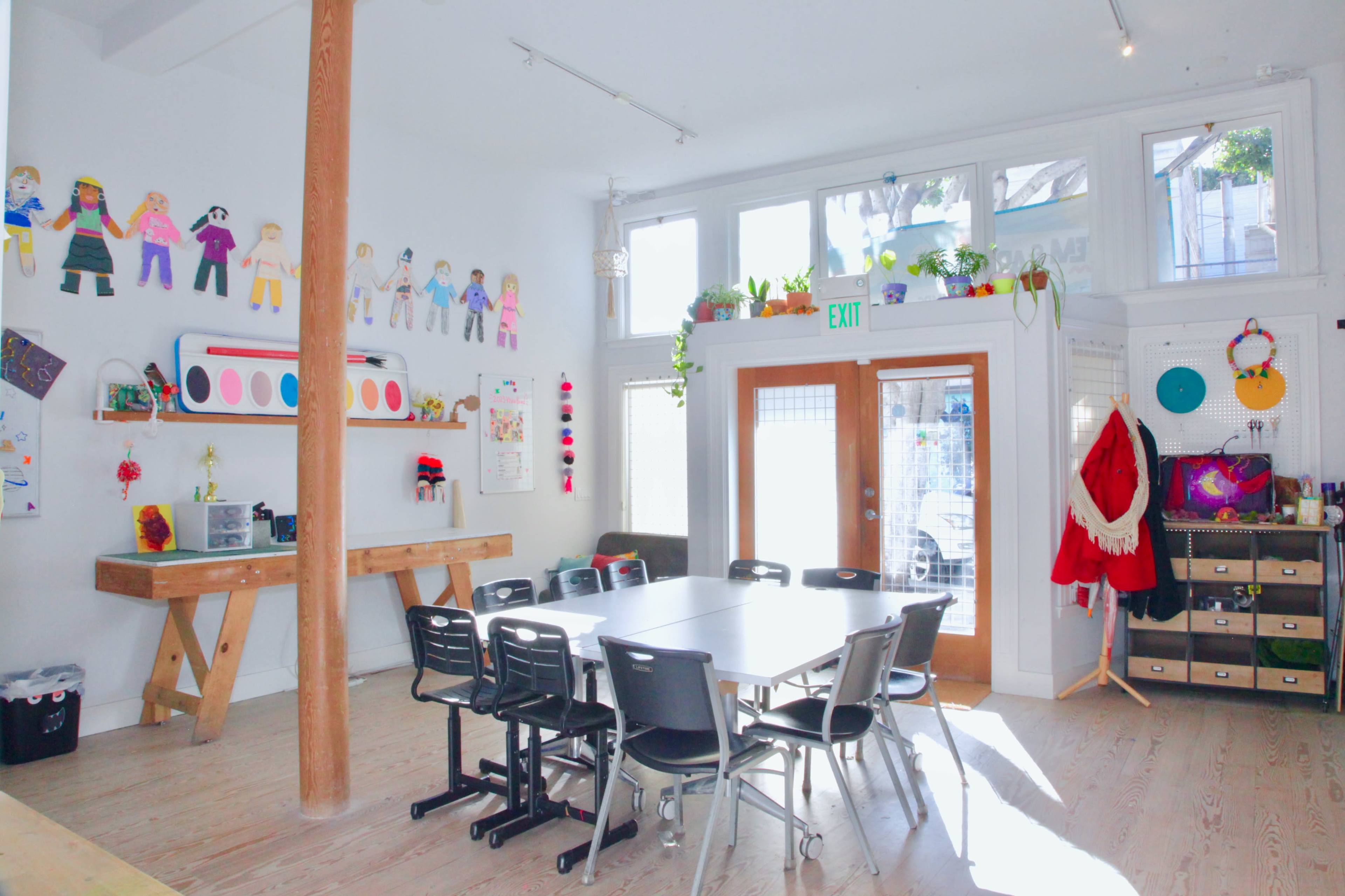 A bright room with a central wooden table surrounded by black chairs, featuring colorful decorations on the walls and large windows letting in natural light.