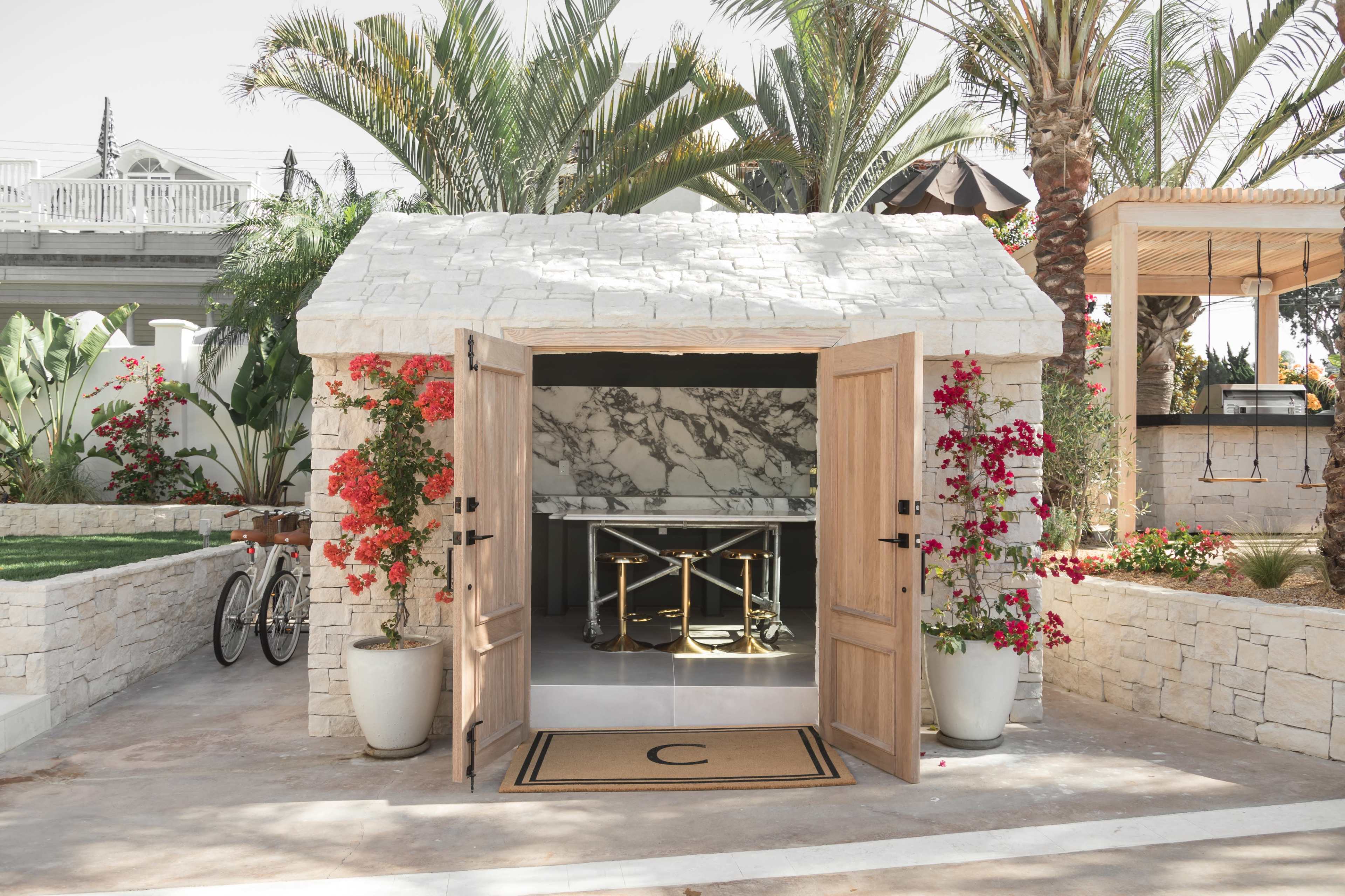 A small stone building with double wooden doors opens to reveal a marble-countered interior, surrounded by vibrant flowers and palm trees.