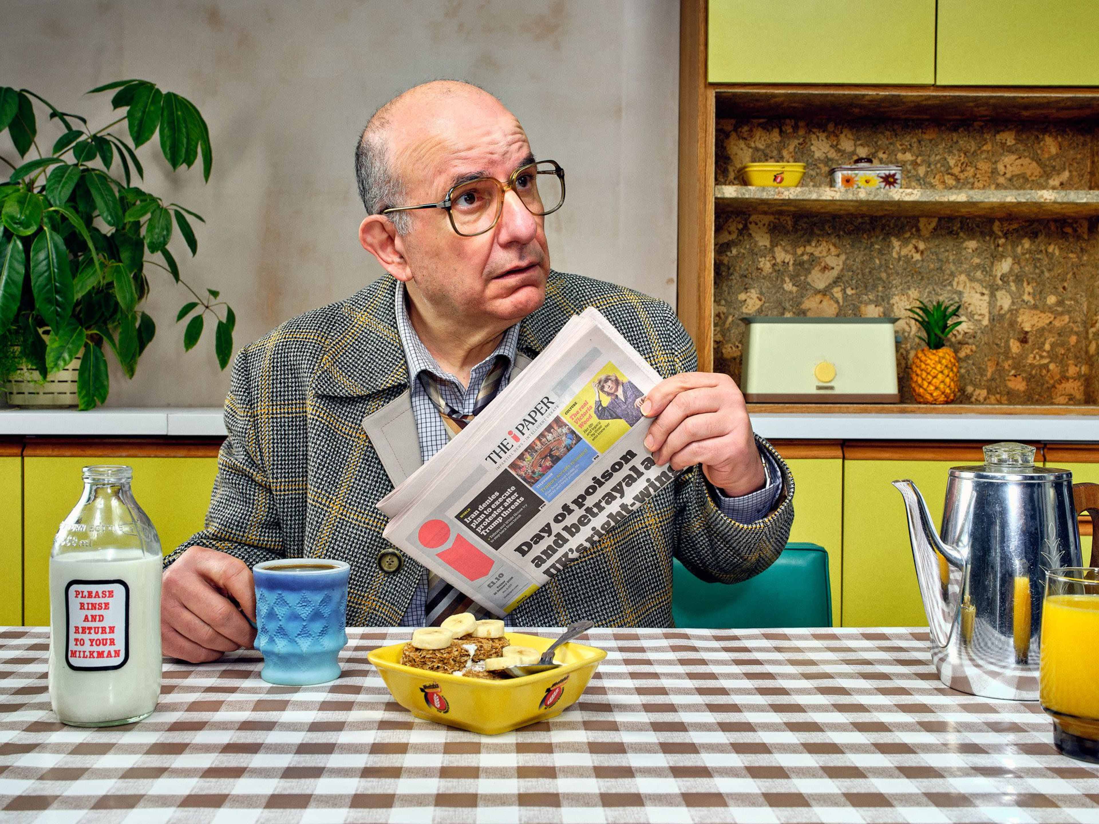 A man sits at a table with a newspaper, a cup of milk, a bowl of cereal, and a teapot in a brightly colored kitchen.
