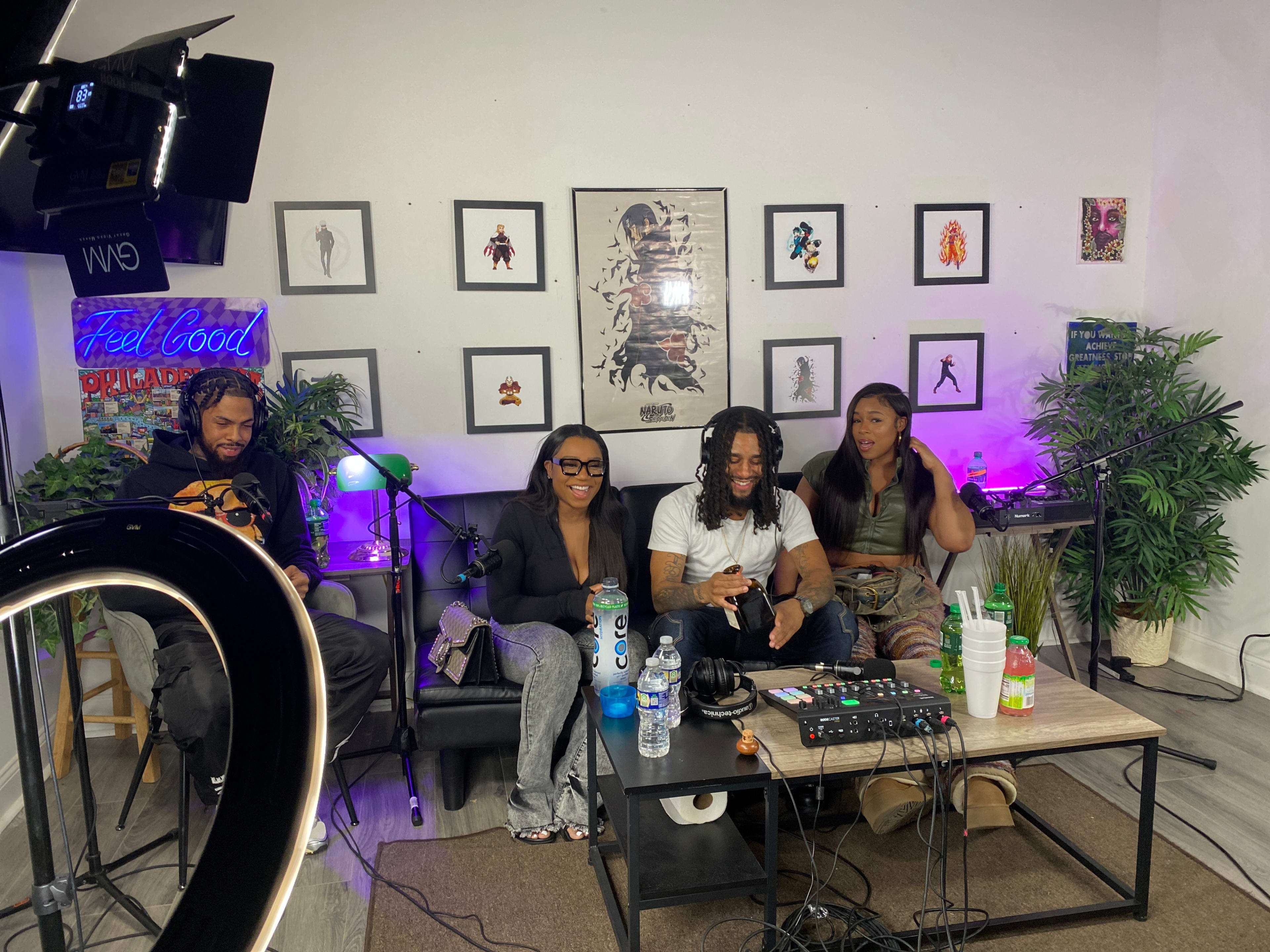 A group of four people are seated on a couch in a studio setting, surrounded by decorations and studio equipment, engaging in conversation.