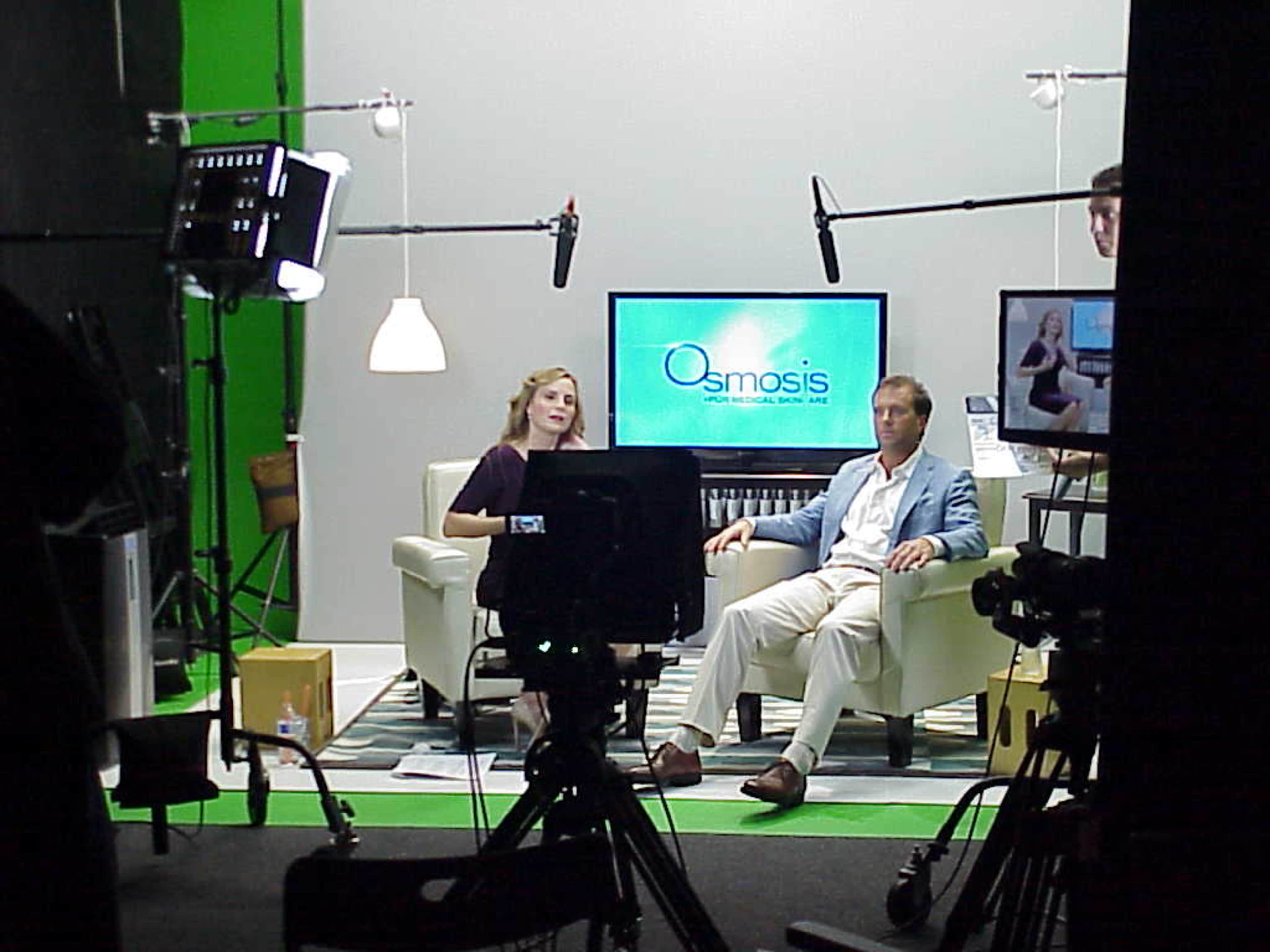 A television studio setup features two presenters seated on a couch with a monitor displaying "Osmosis" in the background, surrounded by cameras and lighting equipment.