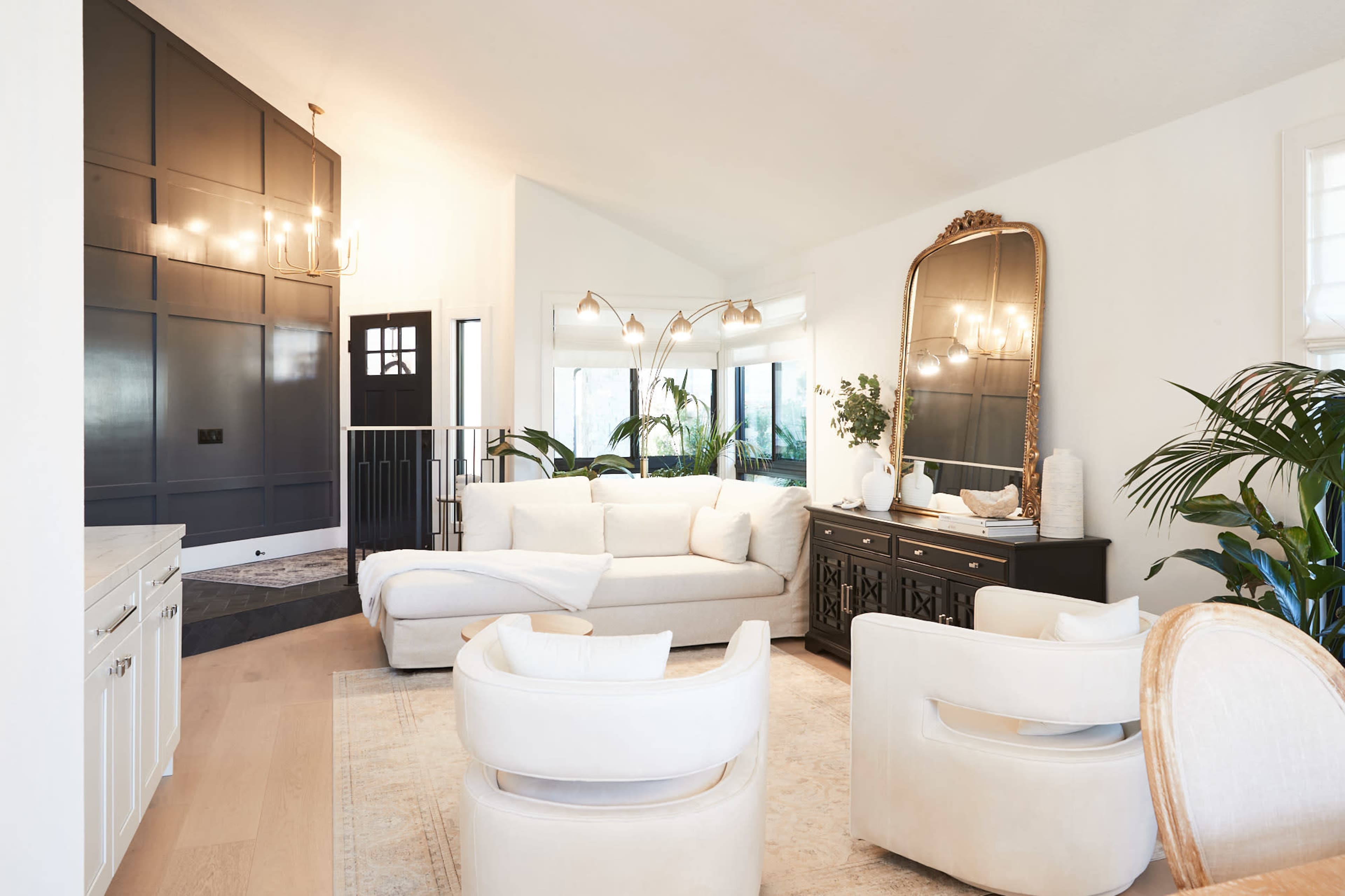 A modern living room featuring a white sectional sofa, two white armchairs, a black and gold mirror, and large windows overlooking greenery.