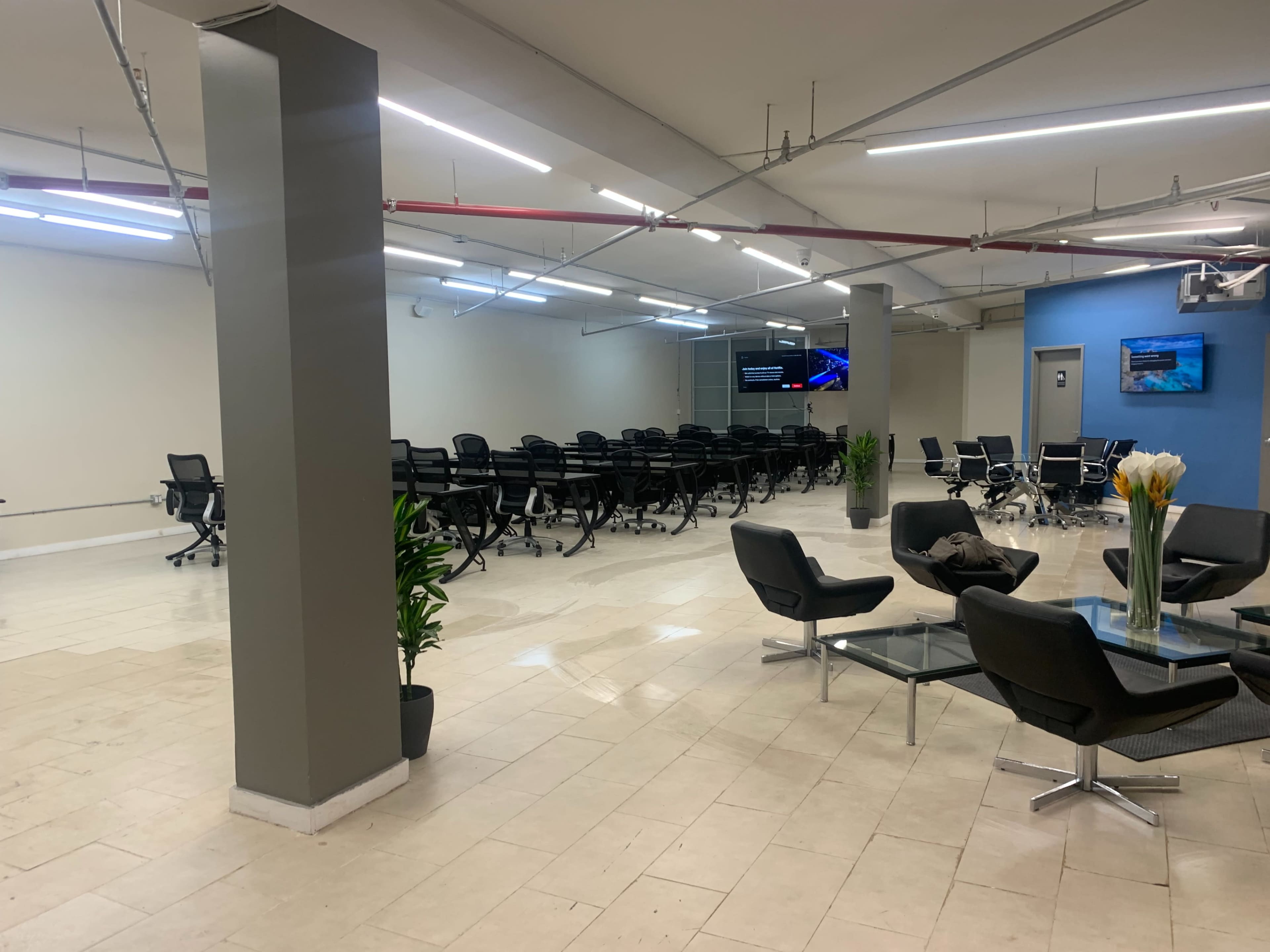 The image shows a spacious room with rows of office chairs positioned in front of a large screen, along with a seating area featuring modern chairs and a glass coffee table.