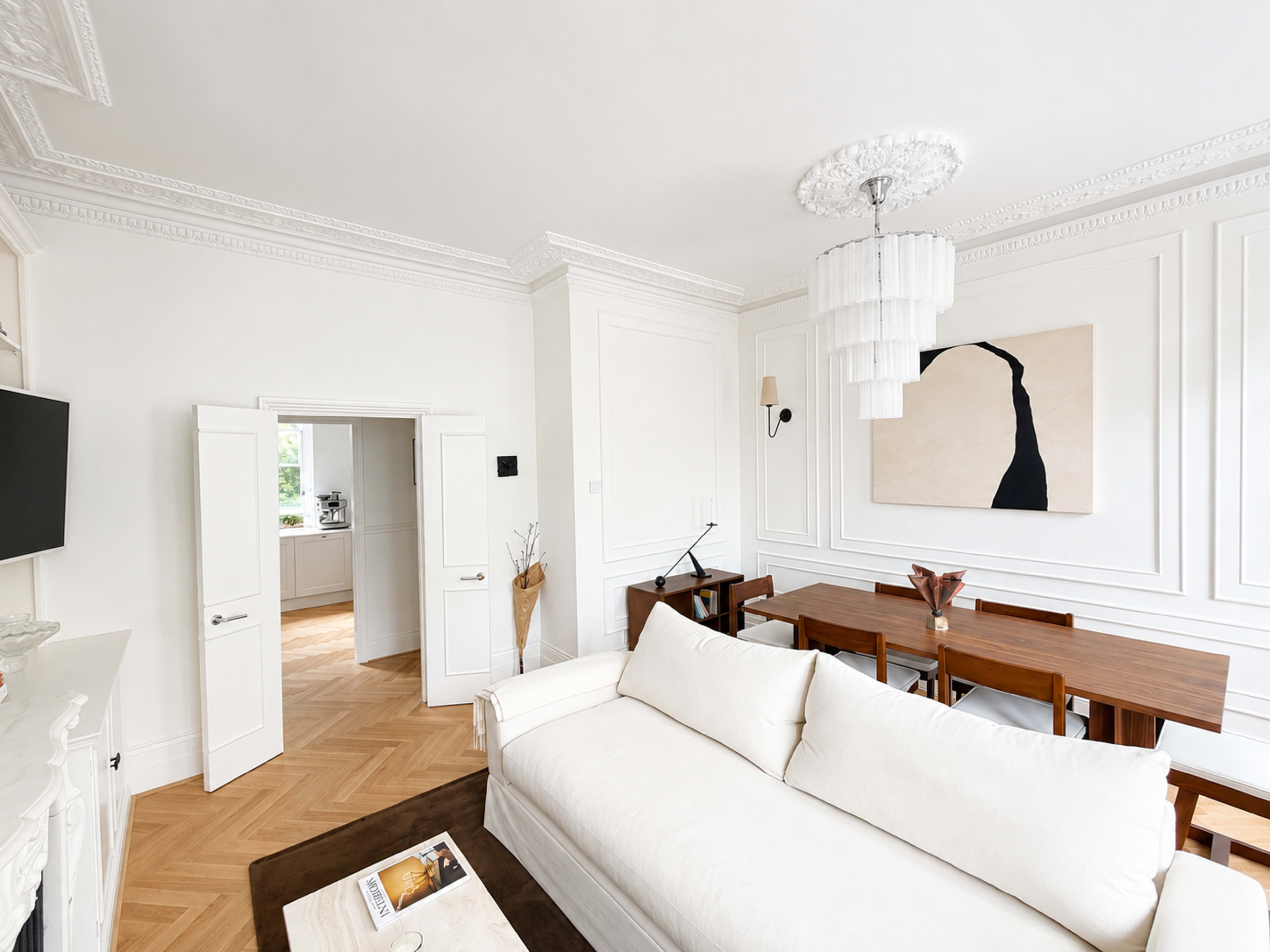 A bright, modern living room features a white sofa, a wooden dining table, and an abstract painting on the wall.