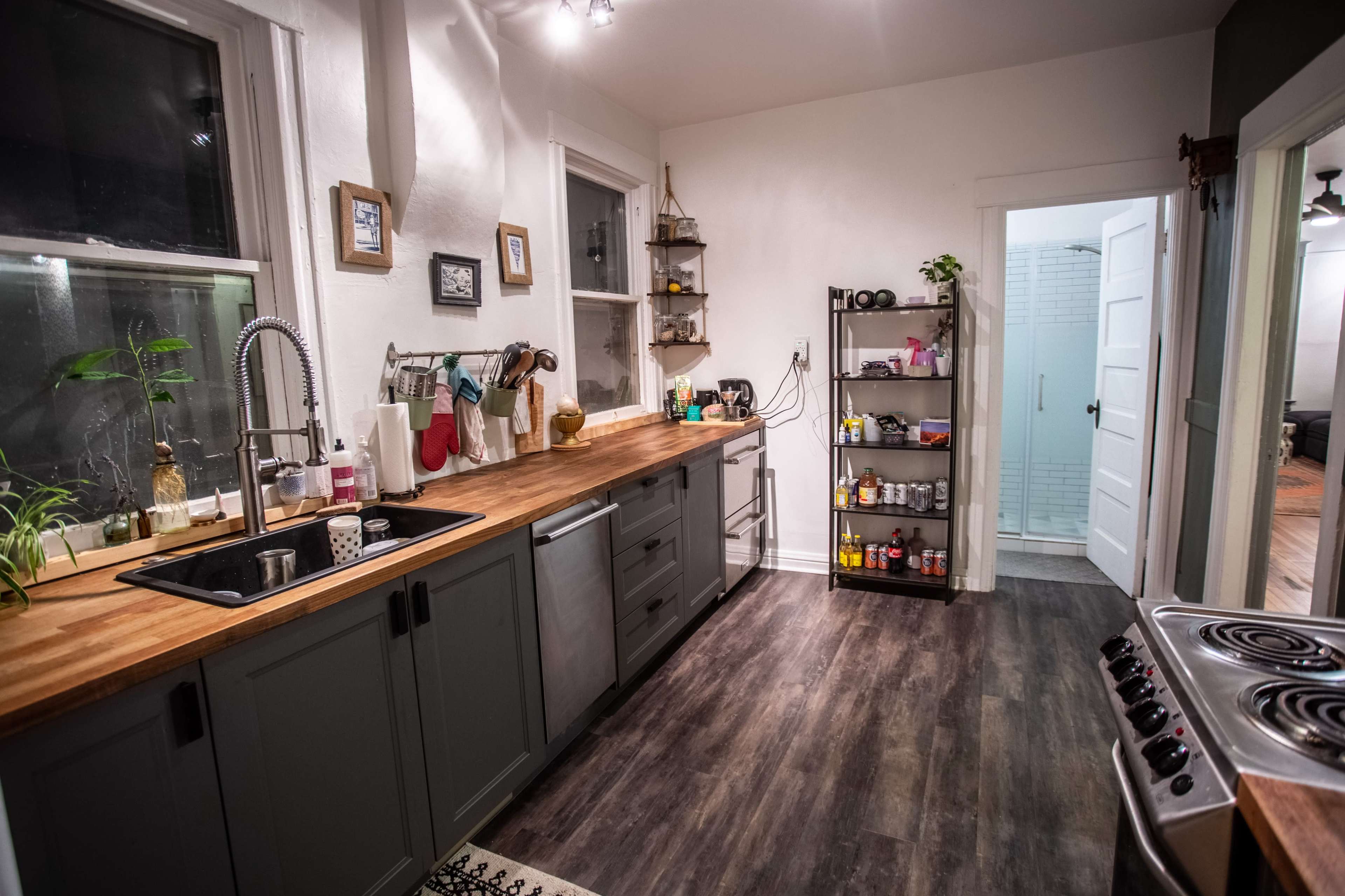 A modern kitchen with dark cabinets, a wooden countertop, a stainless steel sink, and a shelving unit filled with various spices and condiments.