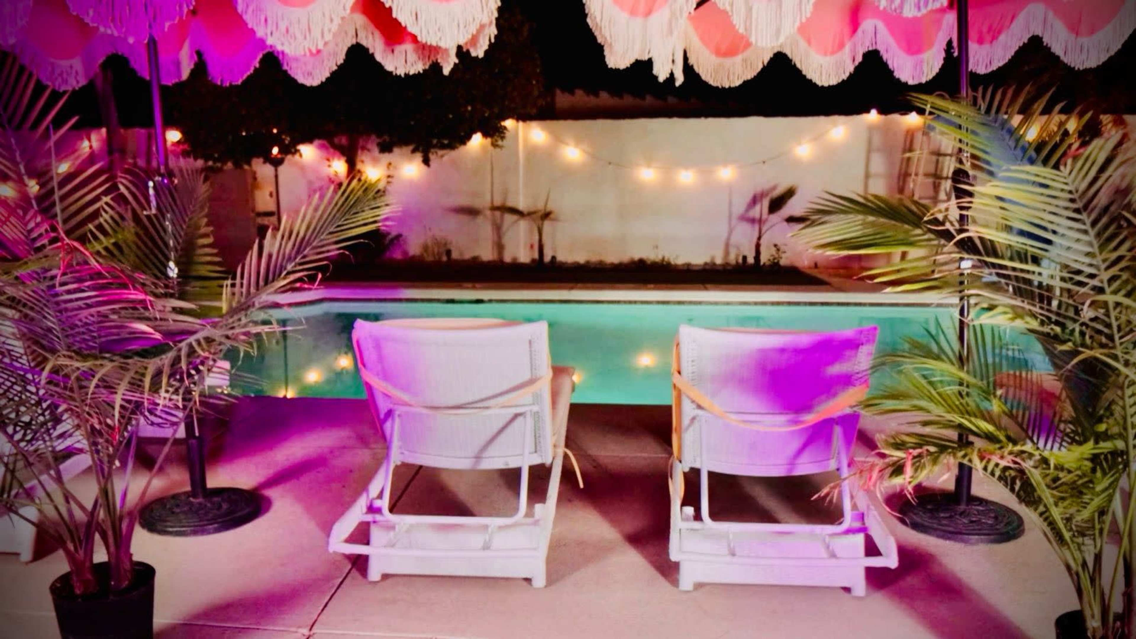 Two white lounge chairs are positioned next to a pool surrounded by palm plants and illuminated by string lights.