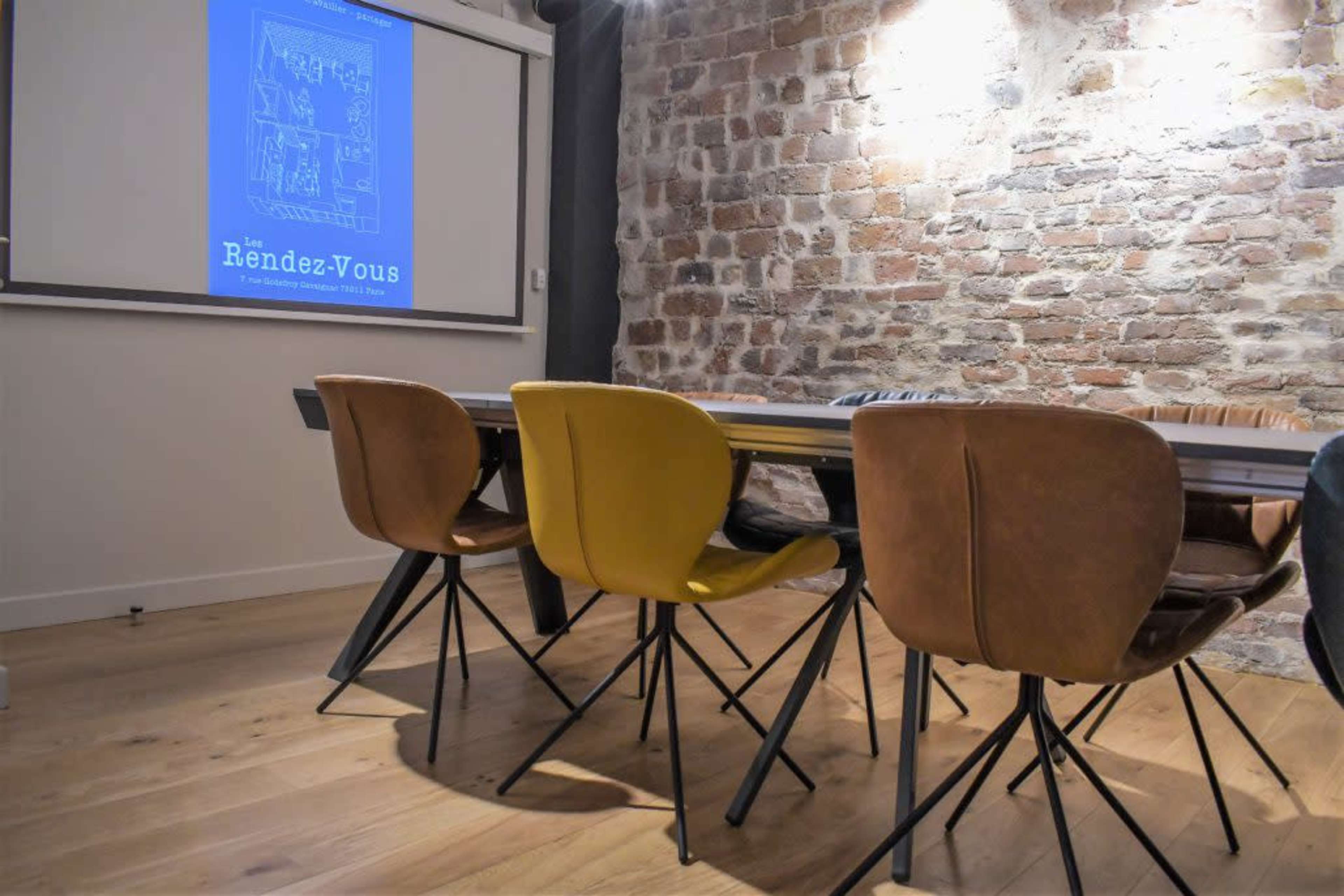 A meeting room features a long table surrounded by chairs, with a presentation slide displayed on a wall behind it.