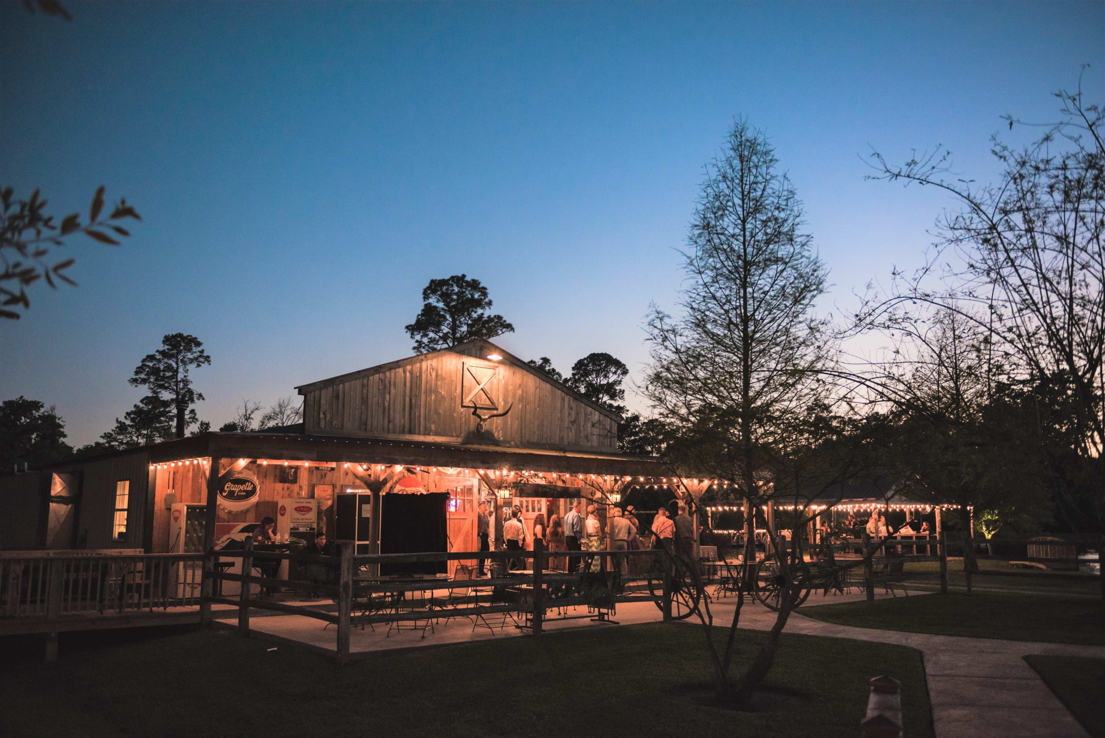 A rustic venue with string lights and a wooden exterior is bustling with people as twilight sets in.