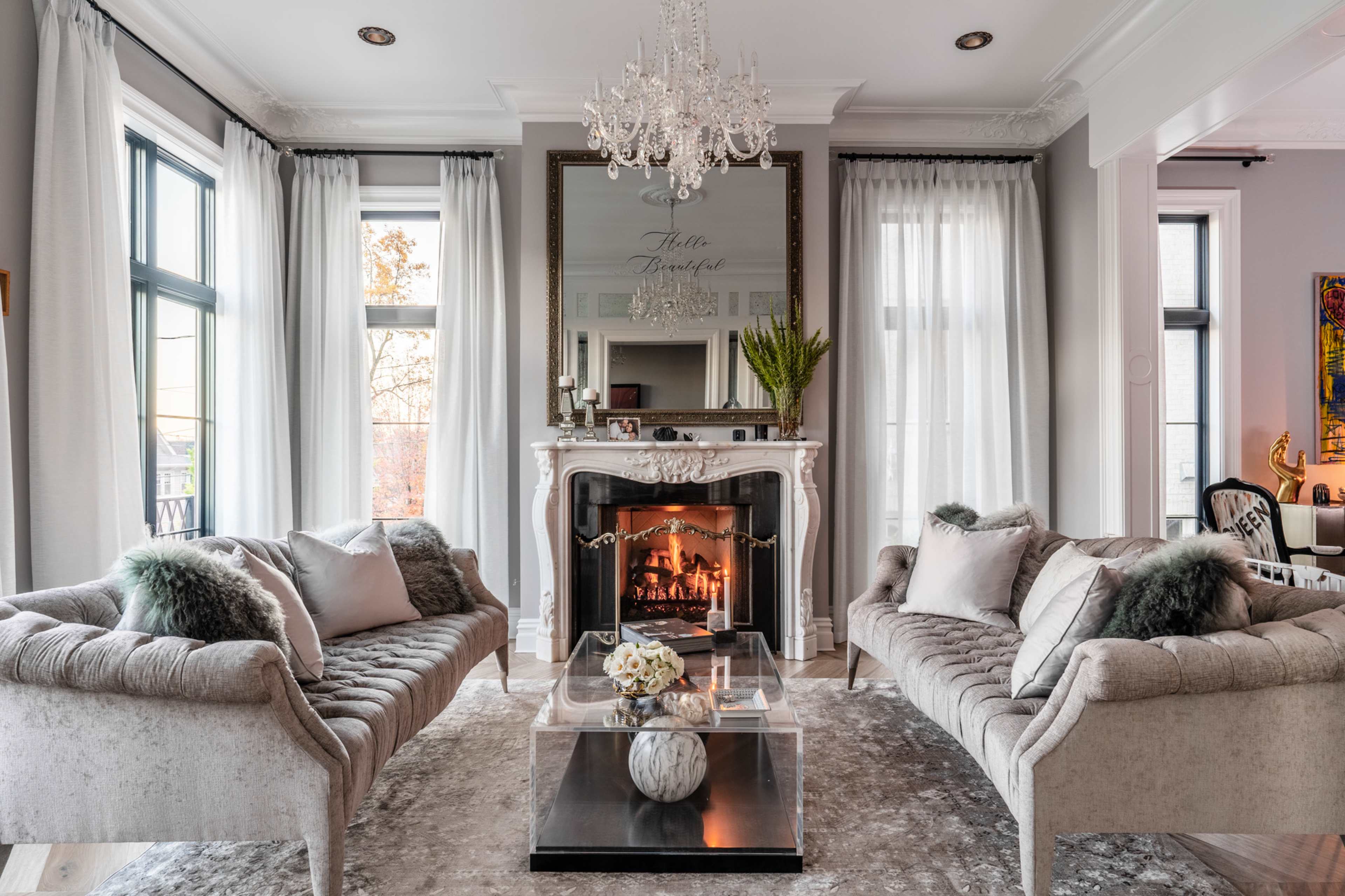 The living room features two elegant sofas facing a decorative fireplace, with a large mirror and a chandelier above, and large windows providing natural light.