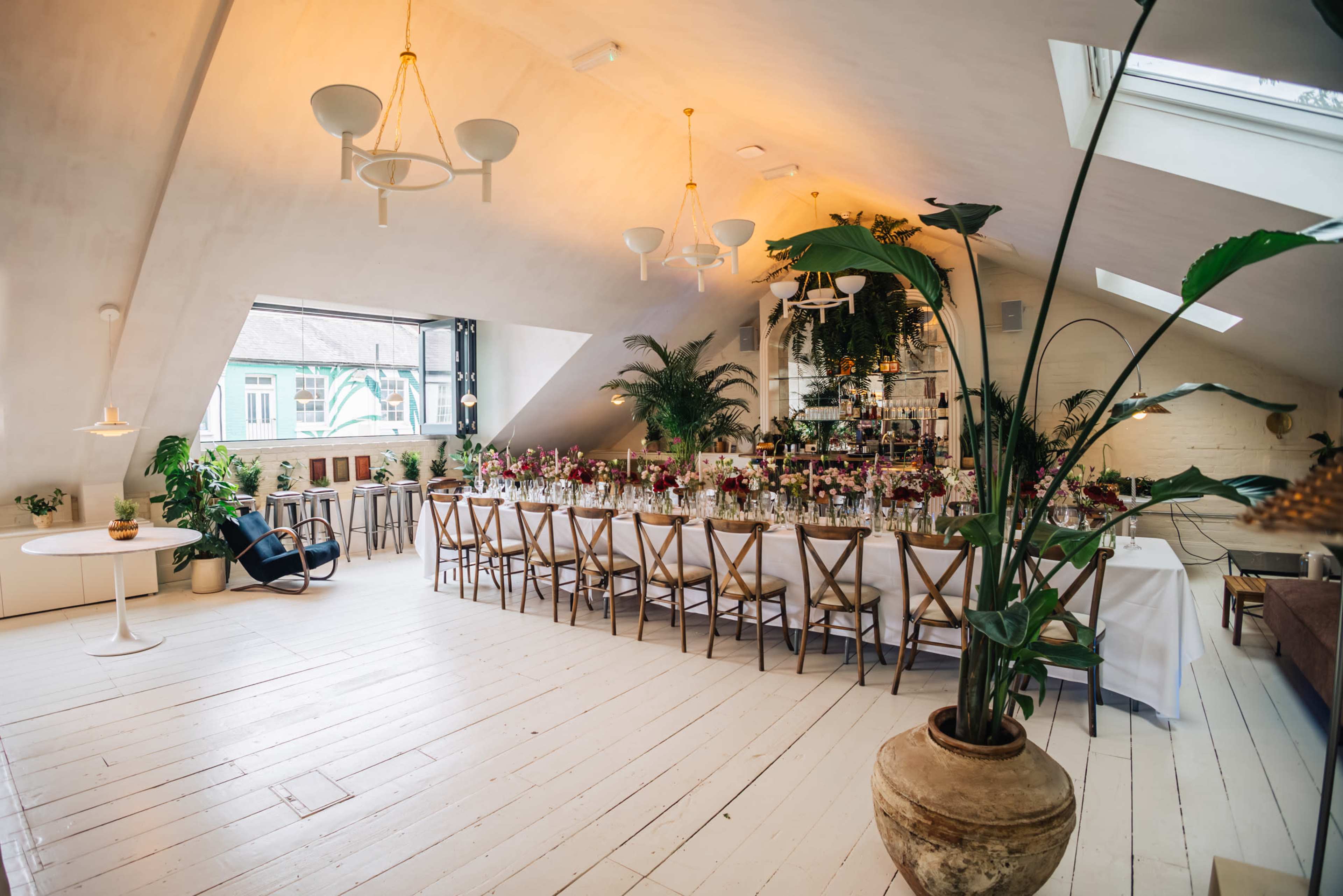 A bright, spacious event room features a long table set for a gathering, surrounded by greenery and modern decor under a sloped ceiling.