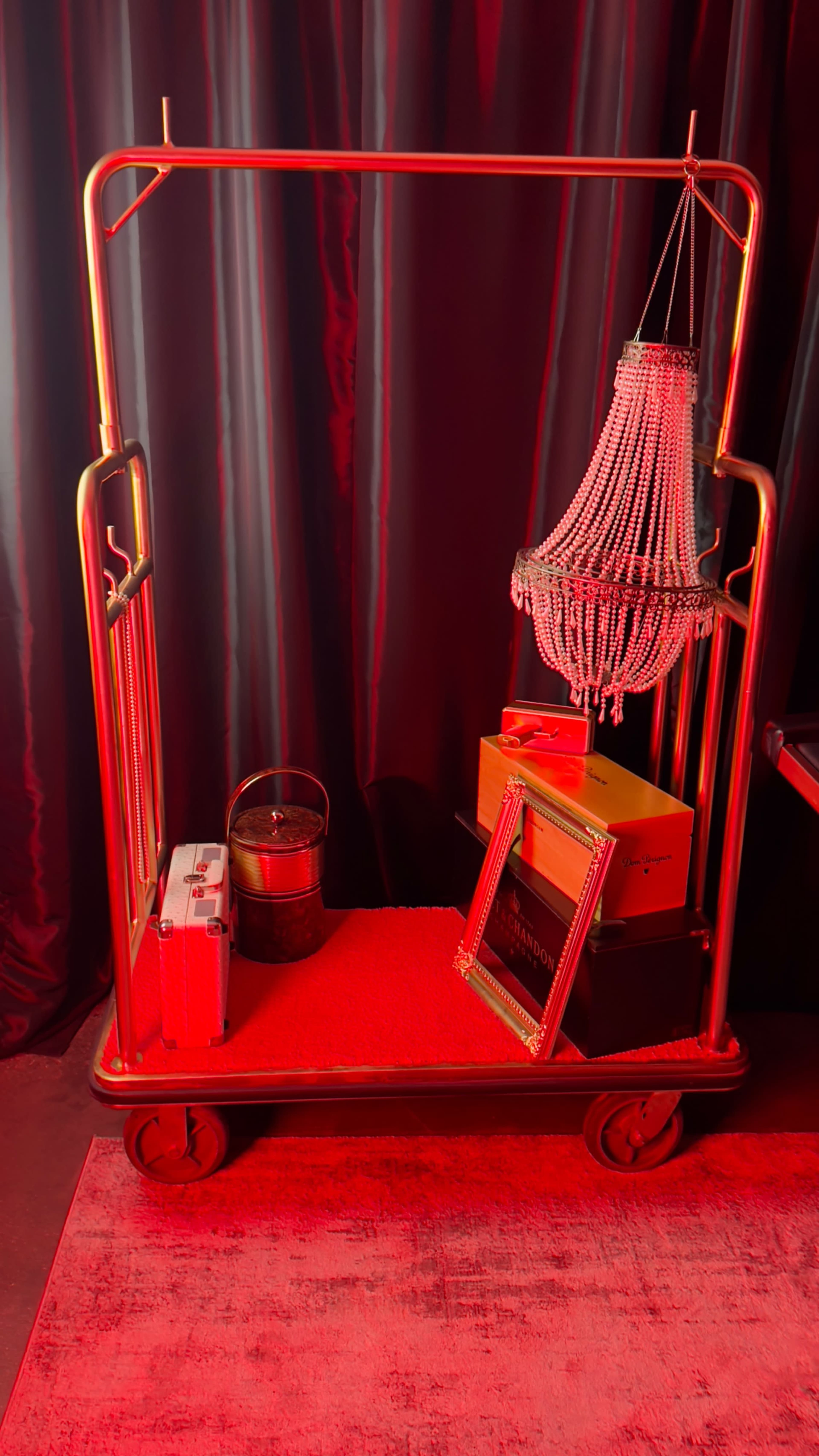 A hotel cart displays a beaded chandelier, various boxes, a suitcase, and a metal bucket against a backdrop of red curtains.