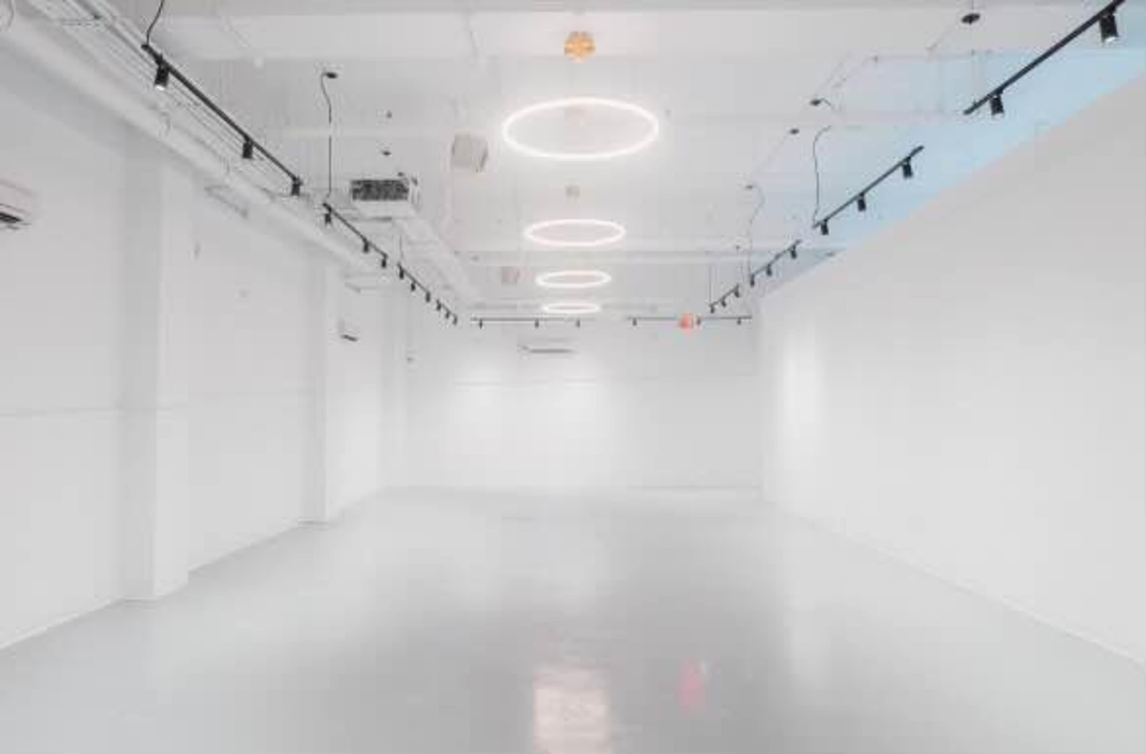 The image shows a spacious, minimalist white room with circular ceiling lights and a polished floor.